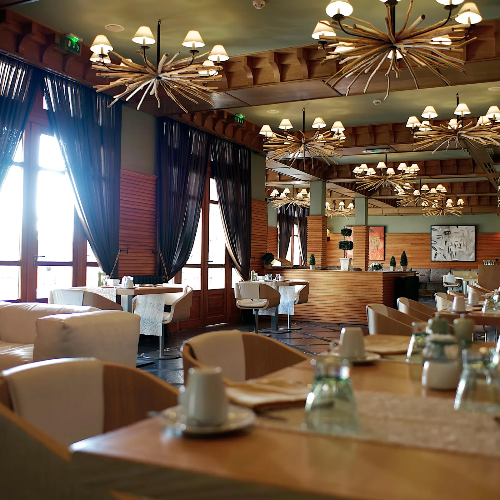 Restaurant intérieur avec des tables dressées, des chandeliers au plafond et de grandes fenêtres drapées de rideaux sombres