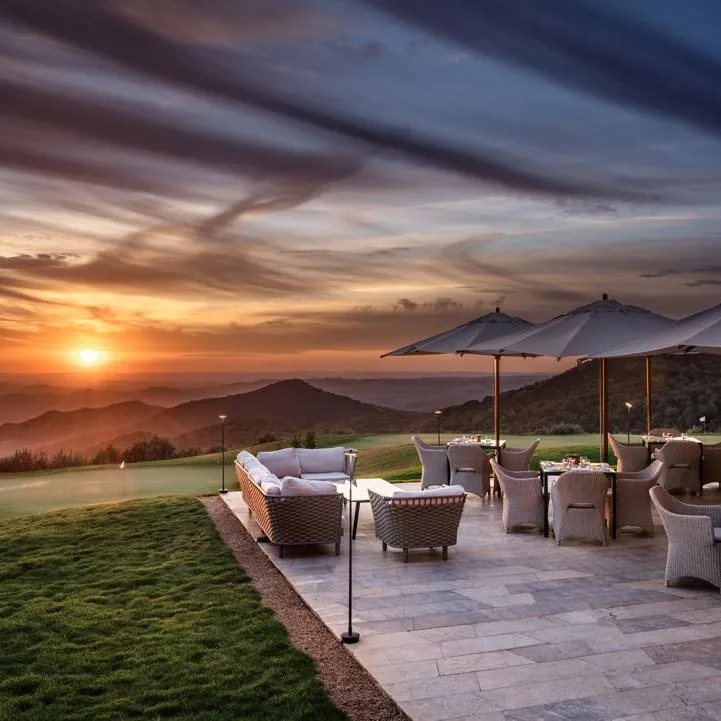 Terrasse avec des chaises et des tables sous des parasols, face à un coucher de soleil sur des montagnes et un terrain de golf