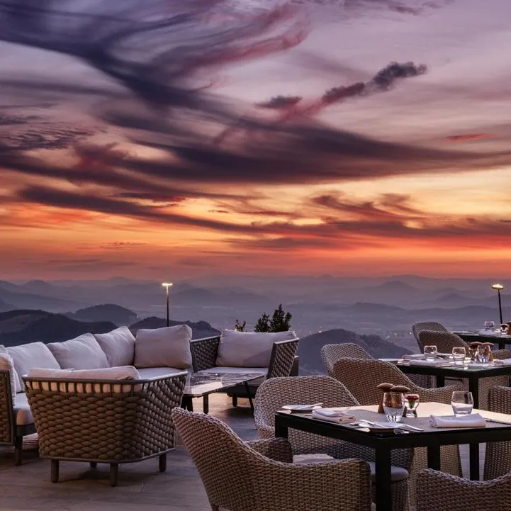 Terrasse avec des tables et des chaises en osier, des coussins blancs et des verres, face à un coucher de soleil sur des montagnes