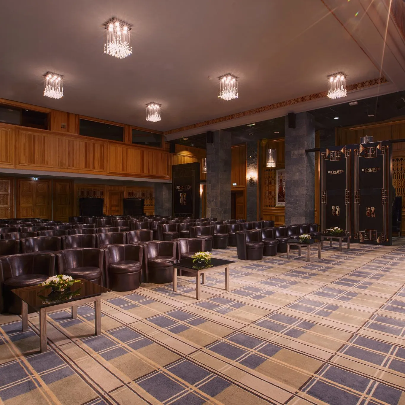 Salle de conférence avec des rangées de fauteuils en cuir marron, des tables basses avec des fleurs, des murs en bois et des lustres au plafond