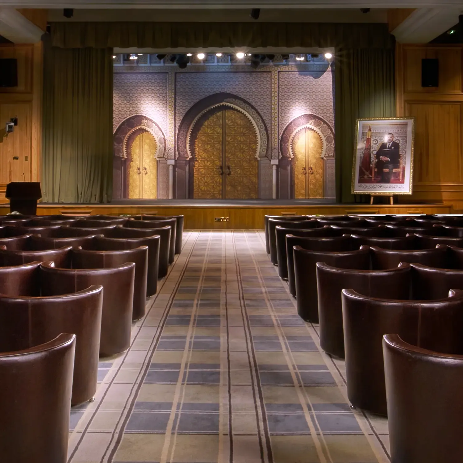 Salle de conférence avec des sièges en cuir disposés en rangées face à une scène ornée de motifs architecturaux et éclairée, avec un portrait sur un chevalet à droite