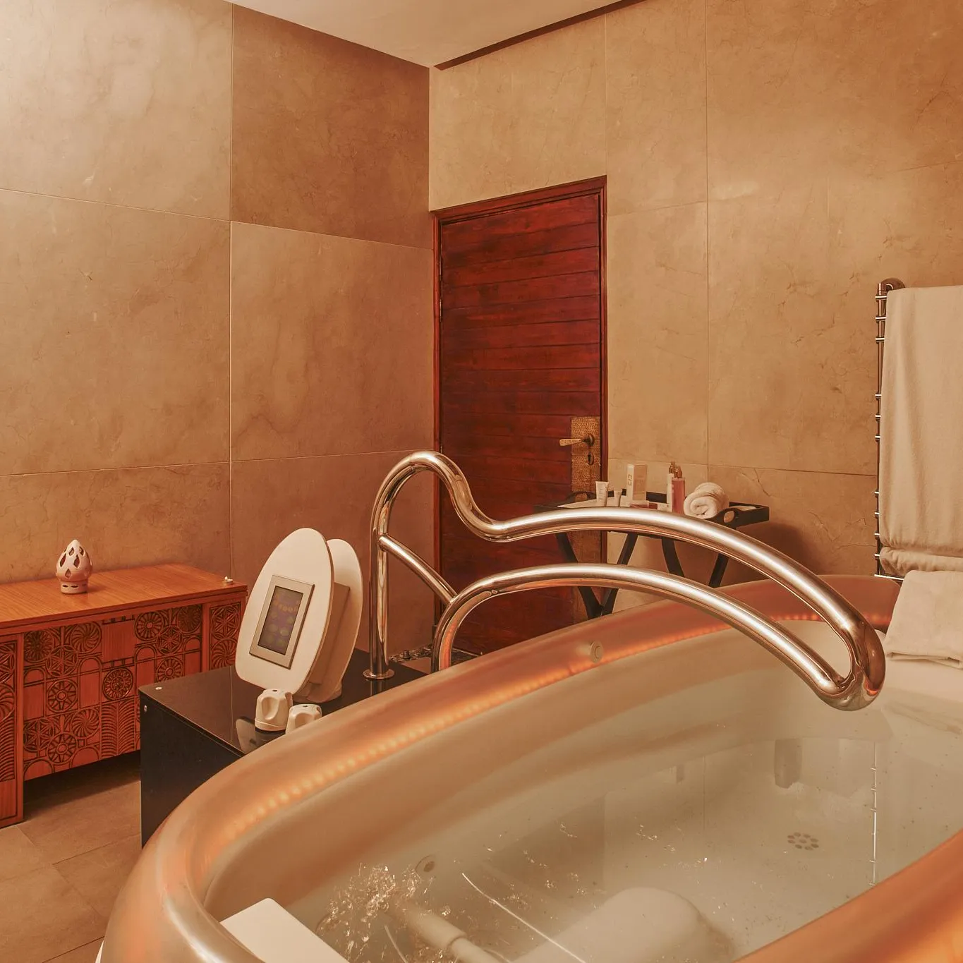 Baignoire remplie d'eau avec des bulles, équipée d'un robinet et d'une pomme de douche, dans une salle de bain aux murs beiges avec un meuble en bois et un porte-serviettes