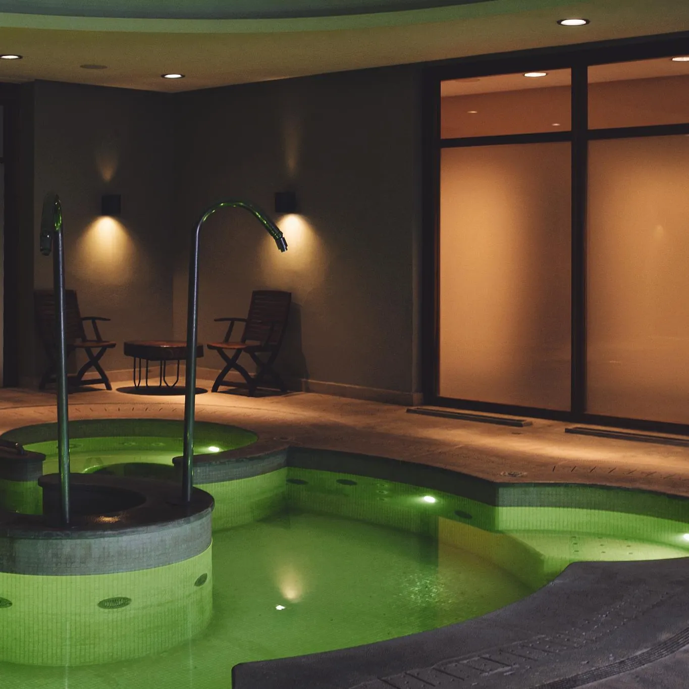 Piscine intérieure éclairée en vert avec deux chaises et une table dans un espace clos aux murs sombres