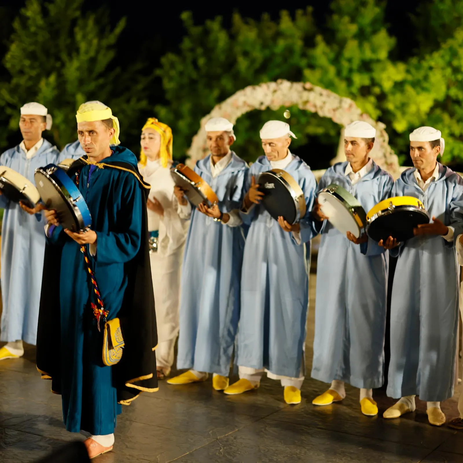 Un groupe de personnes en costumes traditionnels joue de petits tambours sur une scène extérieure décorée de fleurs