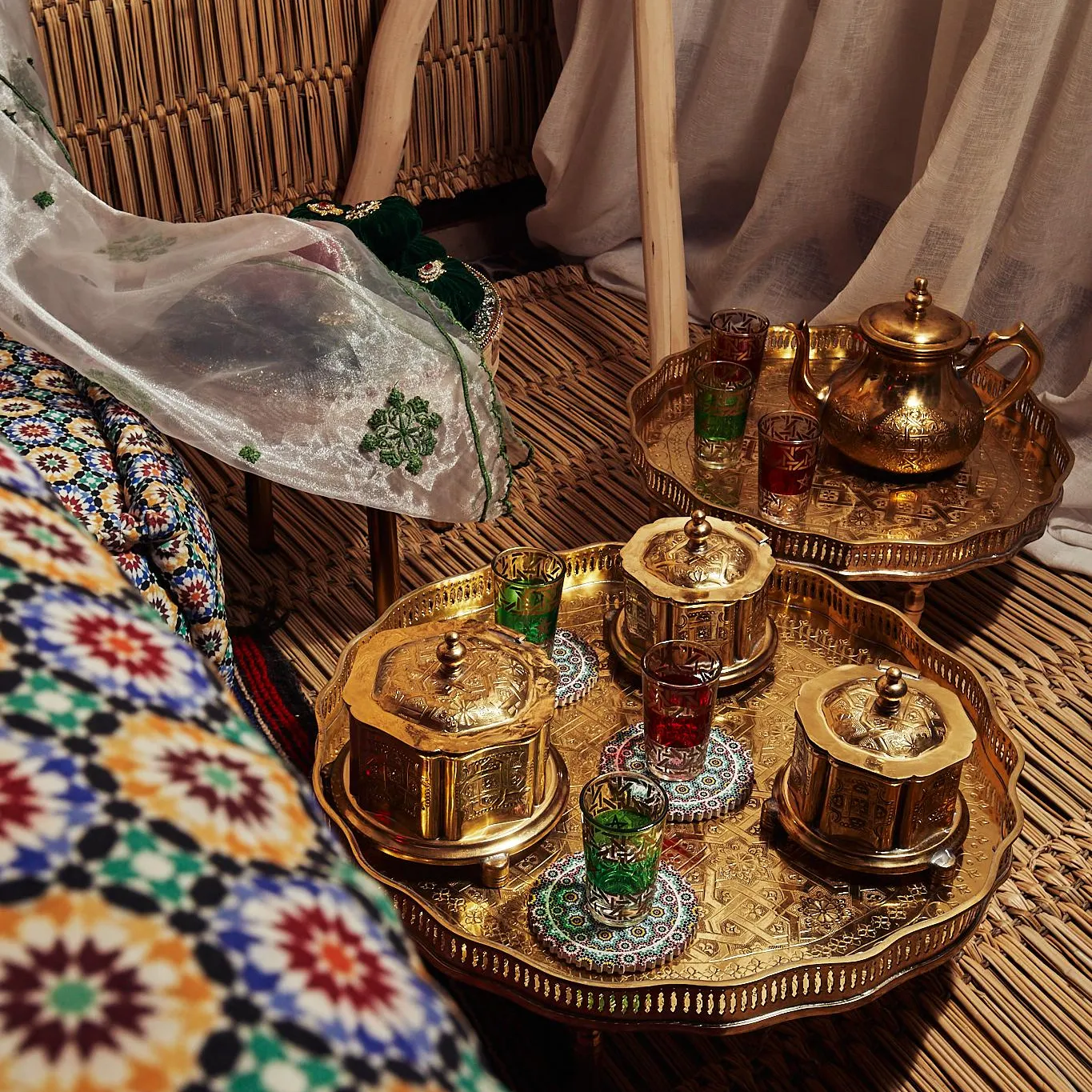Un plateau doré avec des verres et une théière est posé sur une table en bambou, entouré de tissus colorés et de rideaux