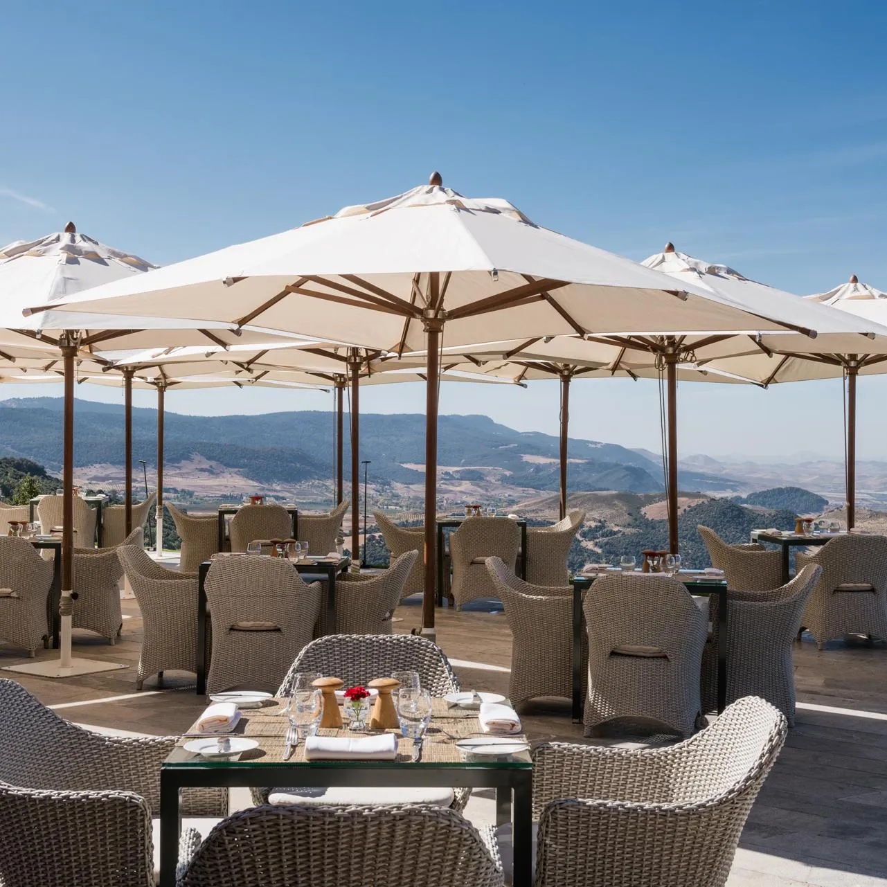 Terrasse de restaurant en plein air avec des tables, des chaises en rotin et de grands parasols, offrant une vue sur des montagnes et une vallée