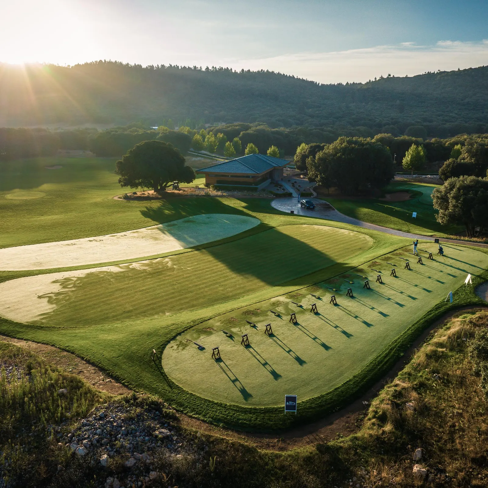Vue aérienne d'un parcours de golf avec des joueurs en action, des bunkers de sable et un club-house en arrière-plan, sous un ciel dégagé