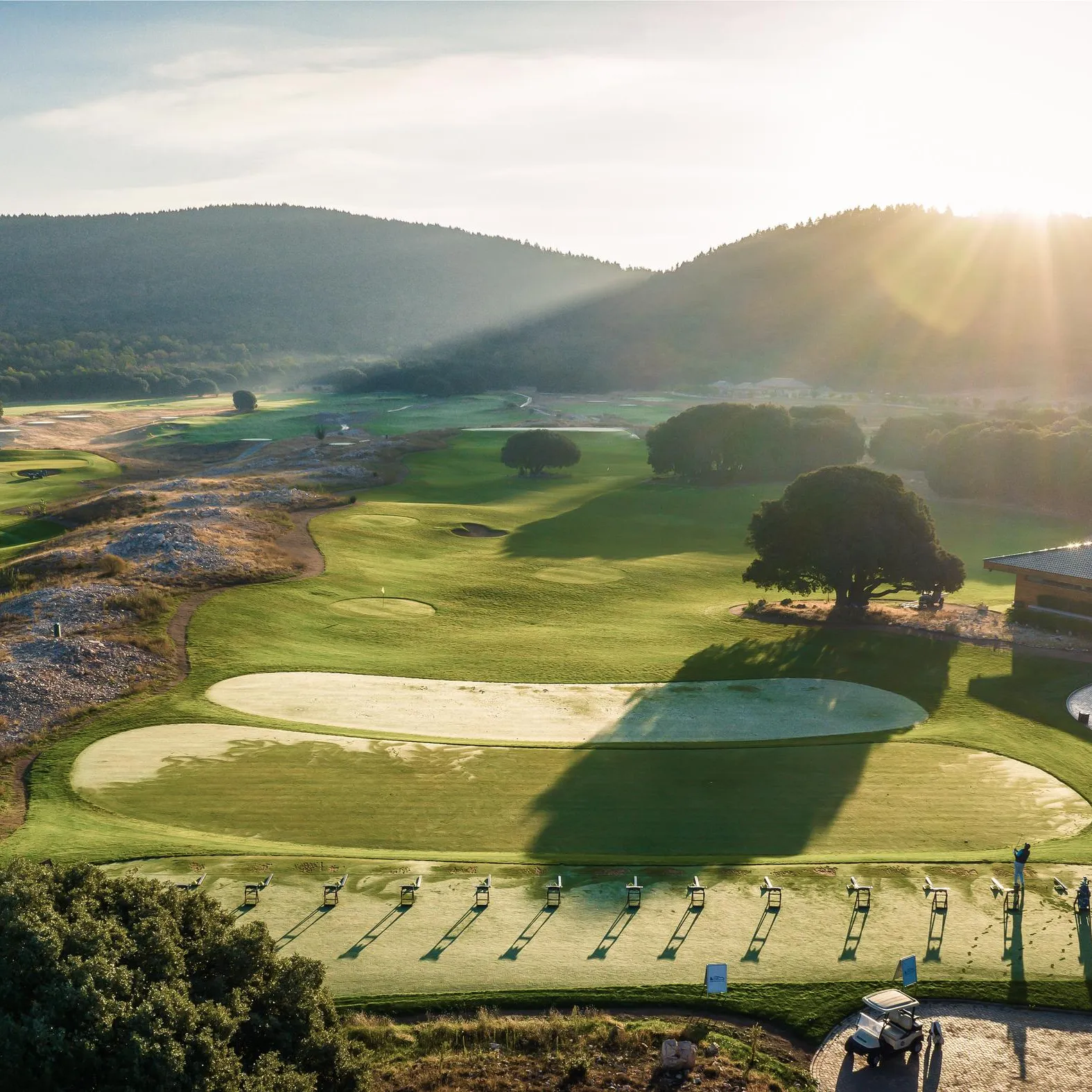 Un terrain de golf avec des practice greens et des cibles, entouré de collines boisées, sous un soleil levant