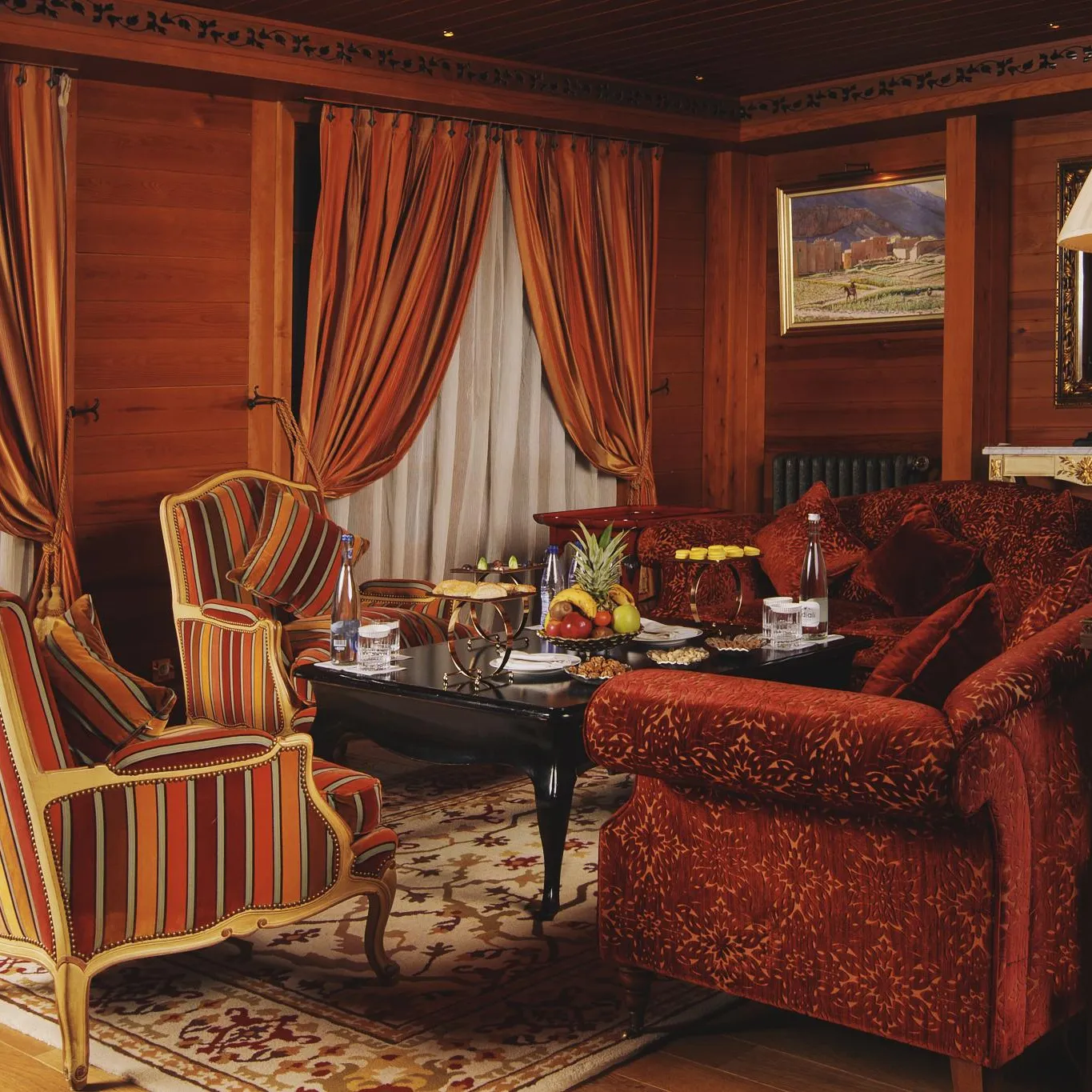 Salon avec des meubles en bois, des fauteuils à rayures et un canapé en velours rouge, décoré de coussins et de tables basses avec des fruits et des boissons