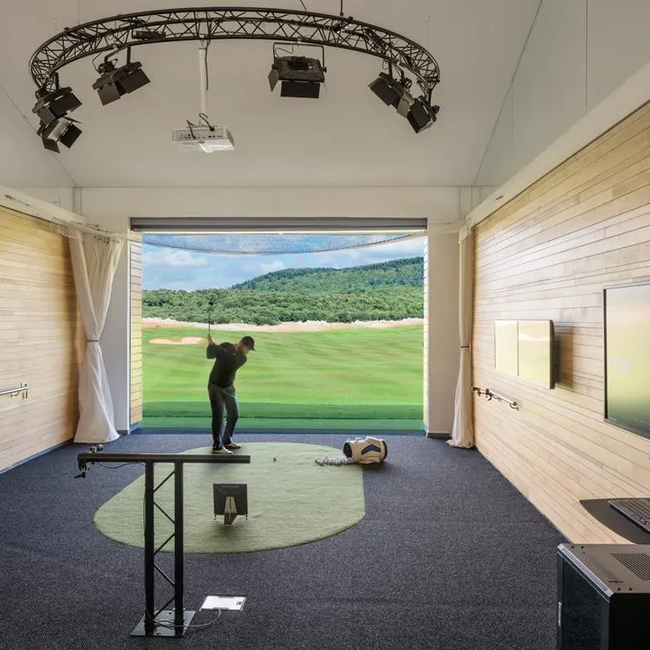 Un golfeur pratique dans un simulateur de golf intérieur avec écran projetant un parcours, équipement et moniteur à côté