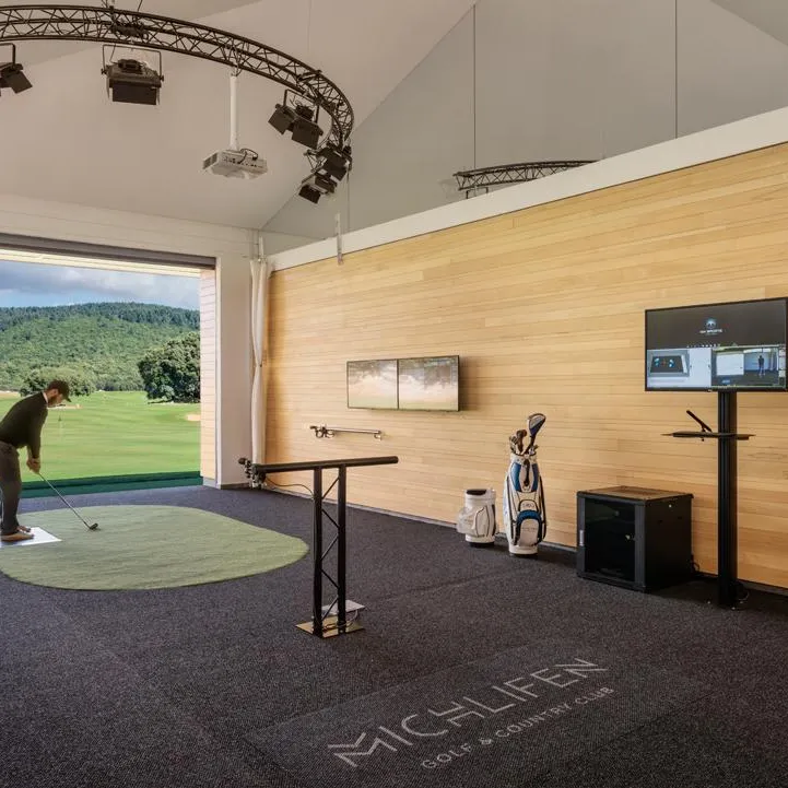 Un golfeur pratique dans une salle d'entraînement intérieure équipée d'un écran projetant un parcours, d'un moniteur et d'un tapis de mise en jeu