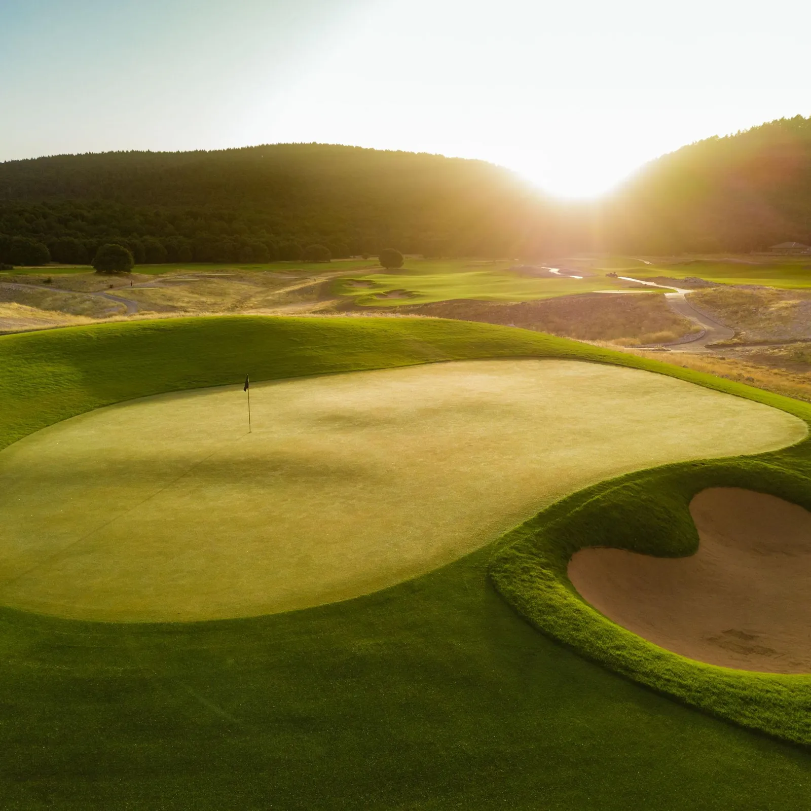 Un green de golf avec un drapeau au centre, entouré de bunkers de sable, sous un soleil couchant avec des collines boisées en arrière-plan