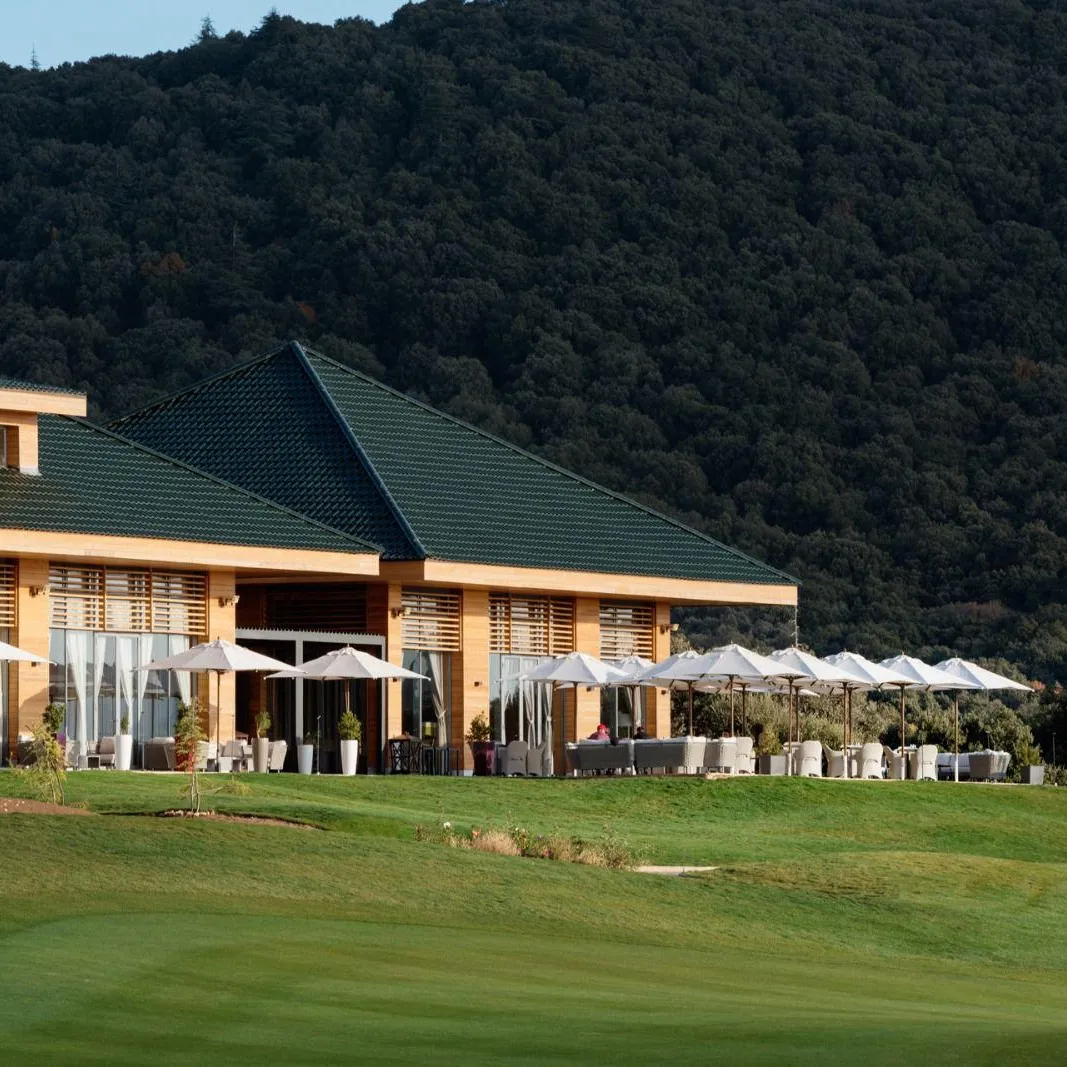 Un bâtiment avec un toit vert et des terrasses extérieures équipées de tables et de chaises sous des parasols, situé près d'un terrain de golf avec une colline boisée en arrière-plan