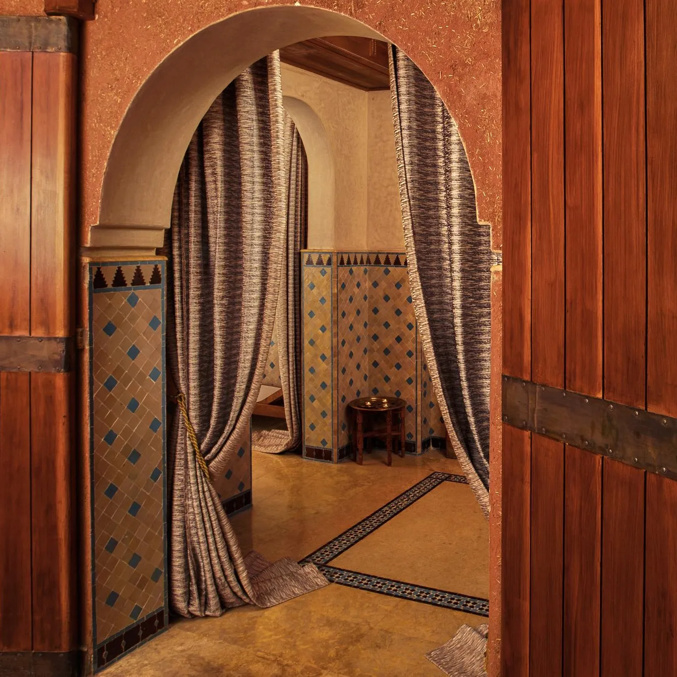 Deux portes en bois avec des anneaux en métal ouvrent sur une pièce aux murs carrelés et aux arches drapées de tissu