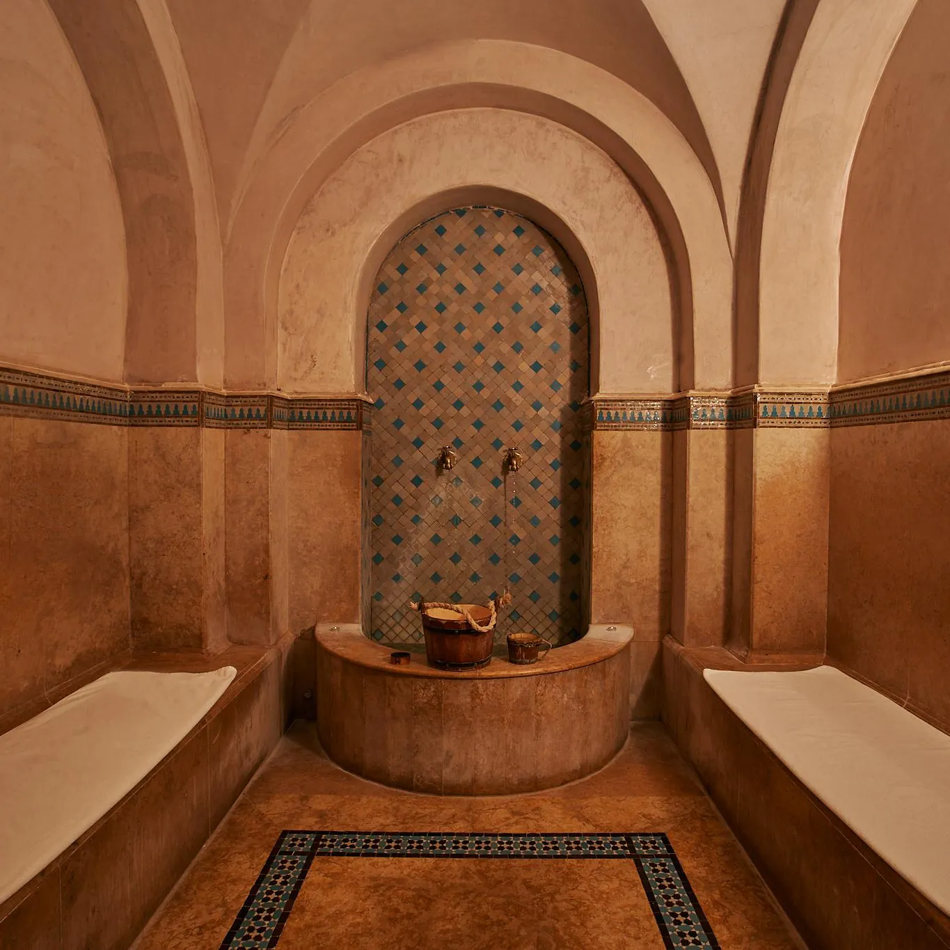 Salle de hammam avec des bancs en pierre, une niche carrelée de motifs géométriques et des seaux en métal