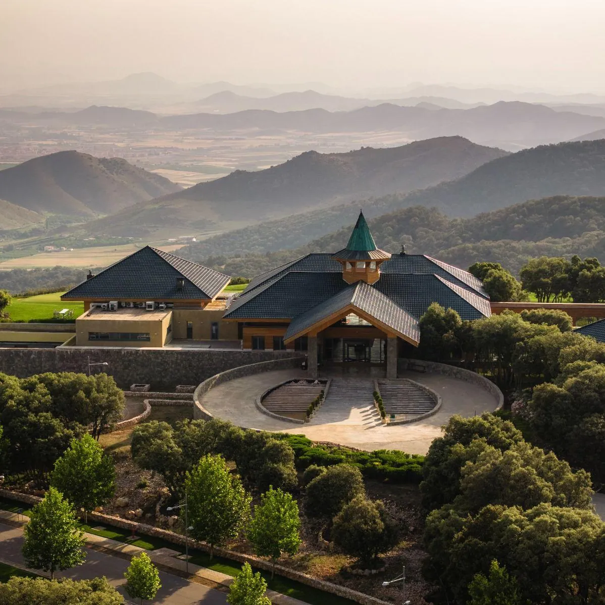 Un bâtiment avec des toits pentus et une tour centrale se trouve dans un paysage vallonné et verdoyant, entouré d'arbres et de jardins