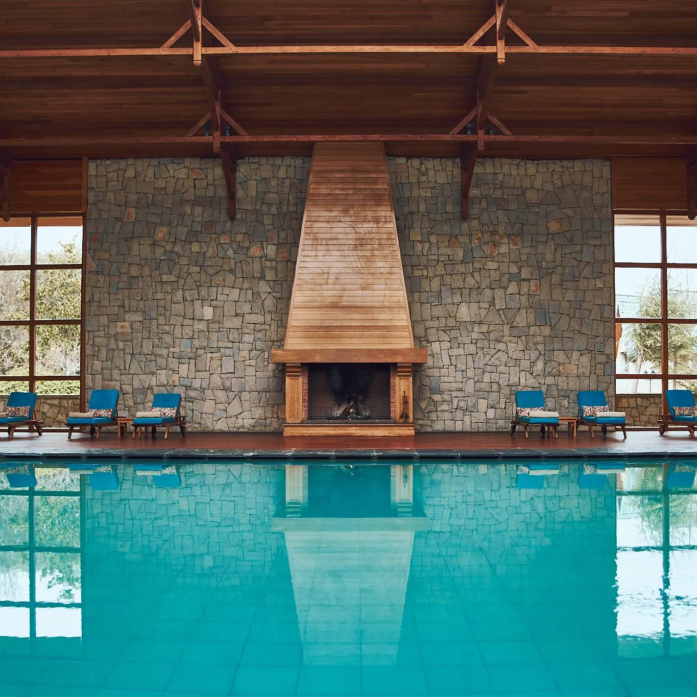 Piscine intérieure avec des chaises longues devant un mur en pierre et une cheminée allumée, entourée de grandes fenêtres en bois