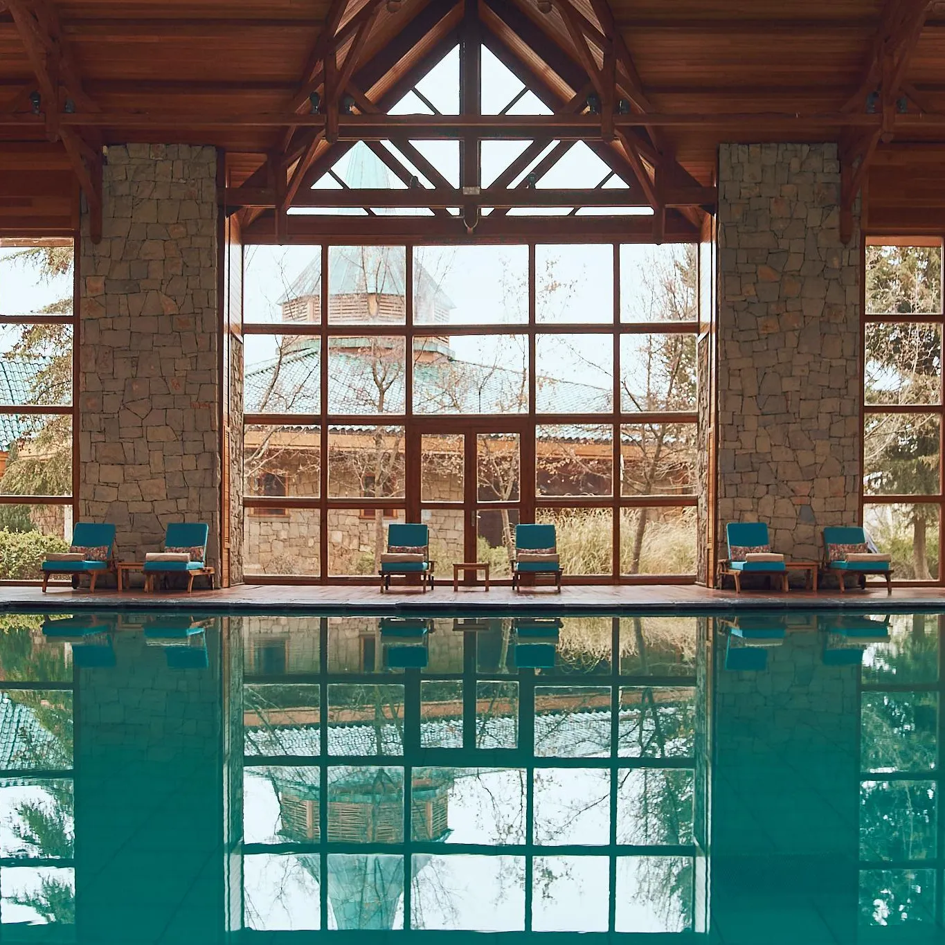 Piscine intérieure avec vue sur des chaises longues et des structures en pierre et bois à travers de grandes fenêtres