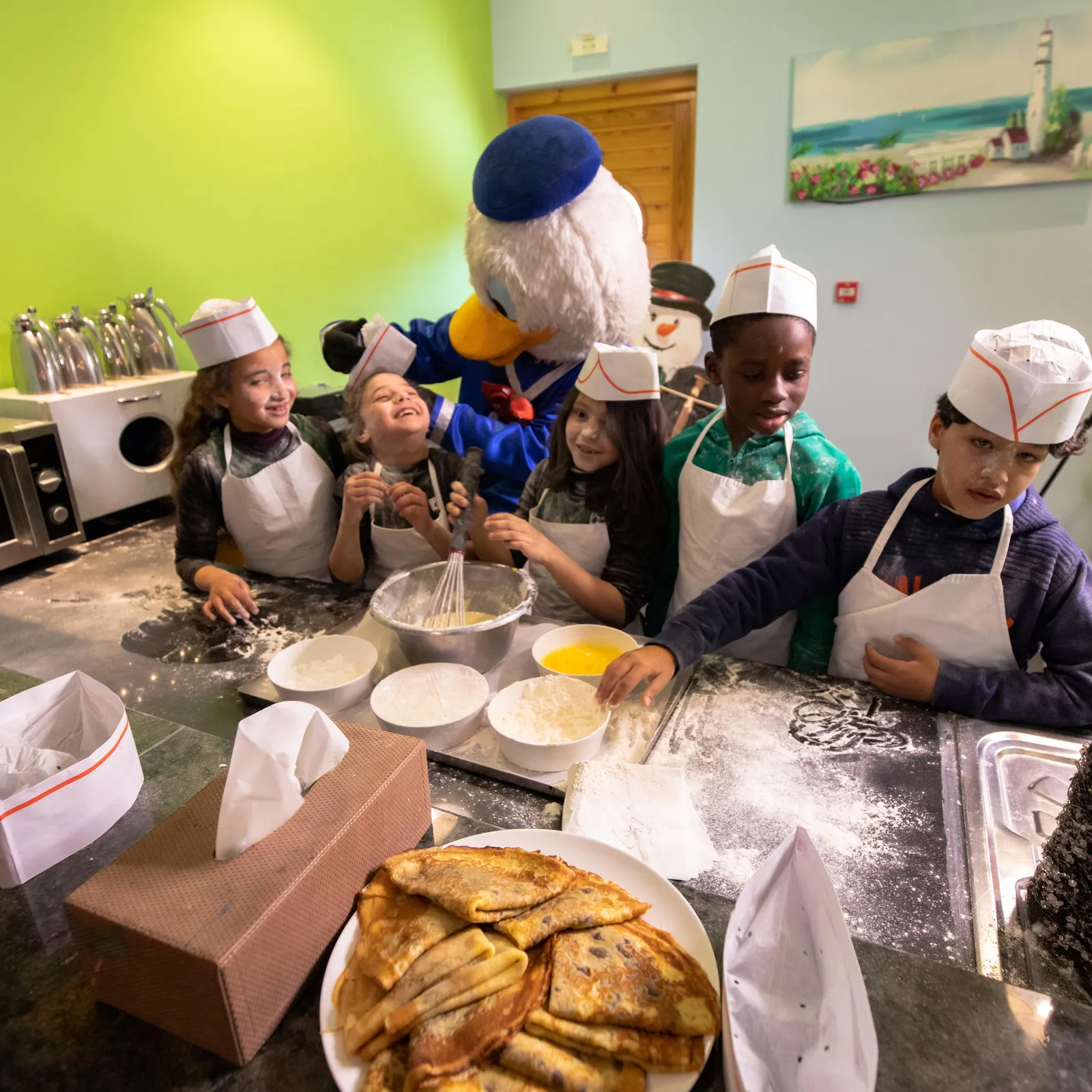 Un groupe d'enfants et un personnage mascottes cuisinent ensemble dans une cuisine, entourés d'ingrédients et d'ustensiles de pâtisserie