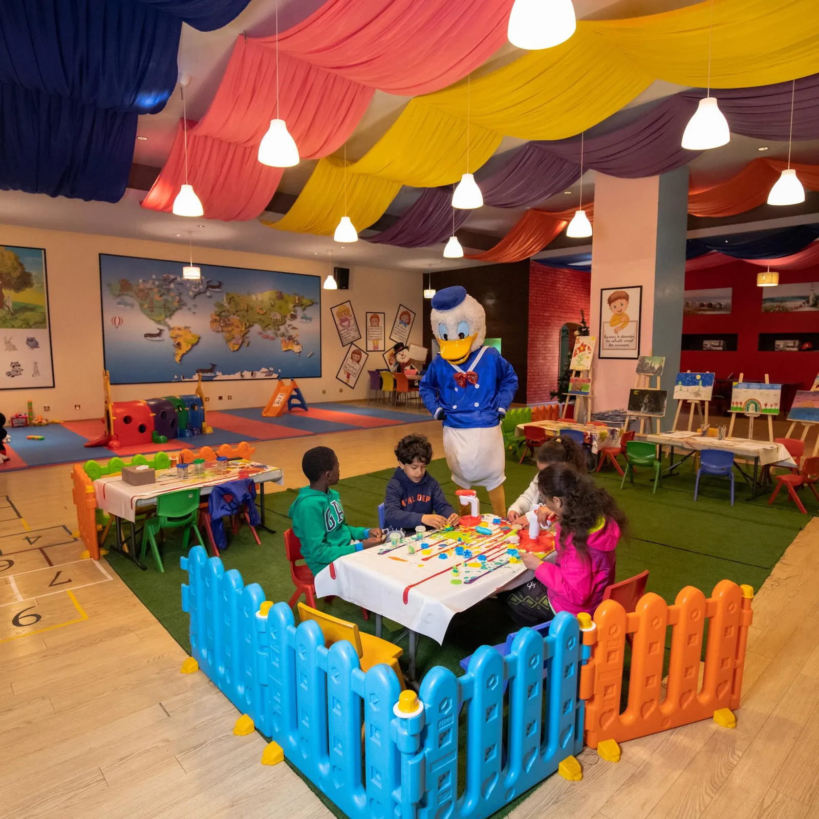 Une salle de jeux colorée avec des enfants assis à des tables, un personnage de dessin animé en costume et des décorations murales éducatives