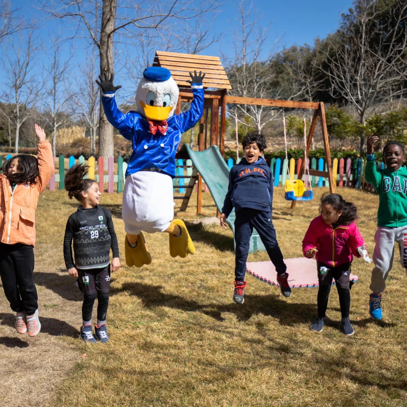 Un personnage costumé en canard saute avec cinq enfants dans un parc avec des jeux en bois et une clôture colorée en arrière-plan