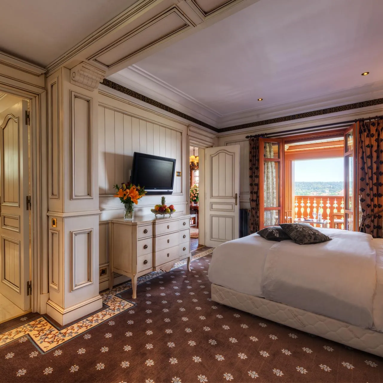 Chambre luxueuse avec lit double, télévision, commode, vue sur balcon et porte ouverte sur salle de bain