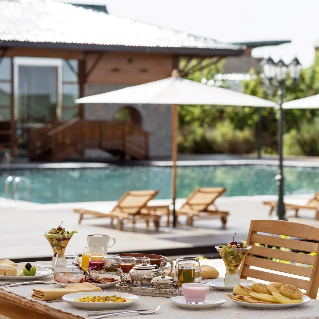 Table de petit-déjeuner en terrasse avec nourriture, boissons et fruits frais, vue sur une piscine avec des transats et parasols