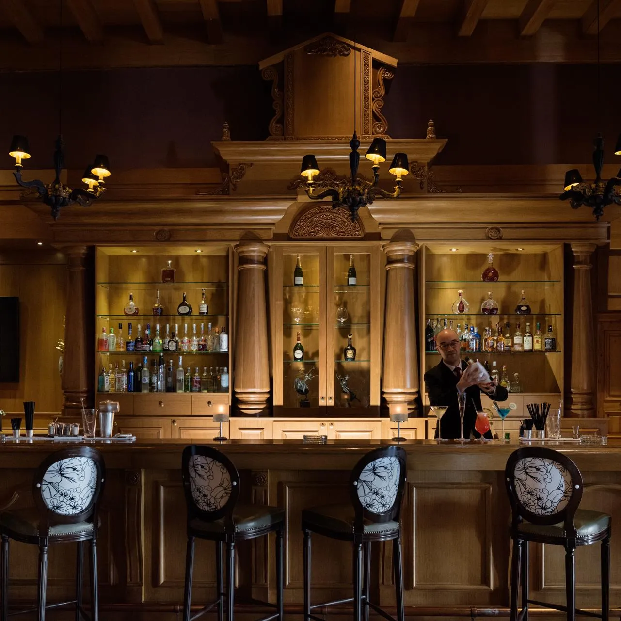 Un barman prépare un cocktail derrière un comptoir en bois avec des étagères remplies de bouteilles et de verres