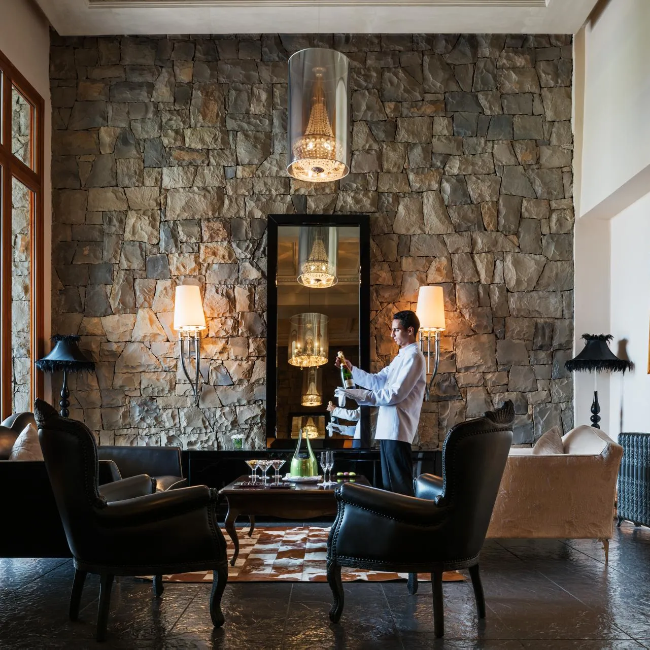 Un serveur en veste blanche prépare des boissons derrière un bar en pierre avec des chandeliers, entouré de fauteuils et de lampes dans un salon élégant
