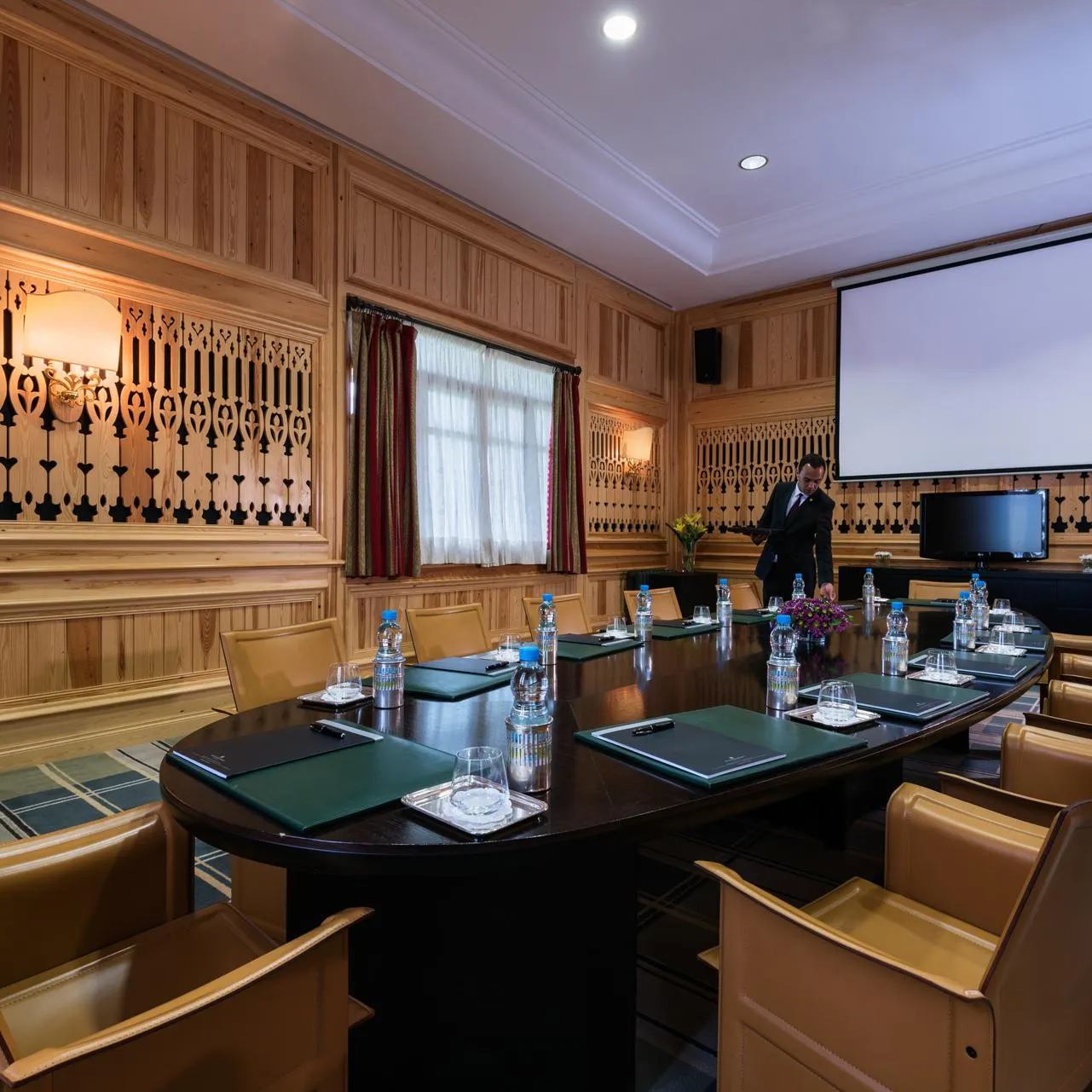 Salle de réunion en bois avec une table ovale, des chaises en cuir, un projecteur et un homme debout près d'un écran