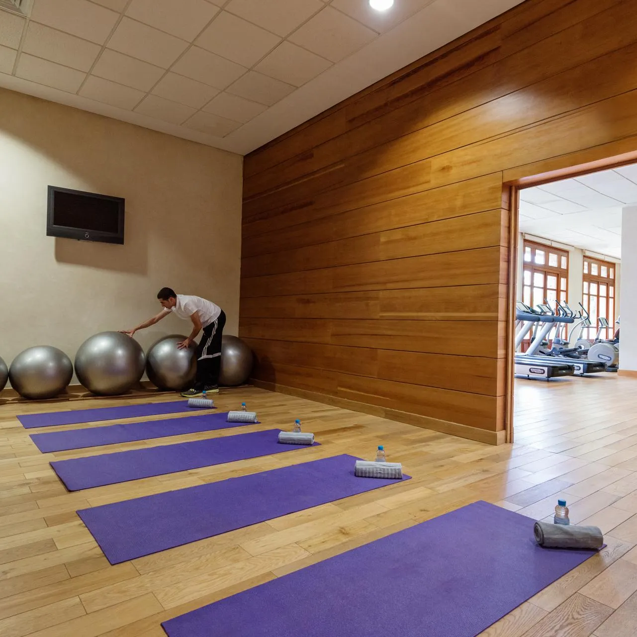 Un homme utilise un ballon d'exercice dans une salle de sport avec des tapis de yoga au sol et des appareils de fitness visibles à travers une porte ouverte