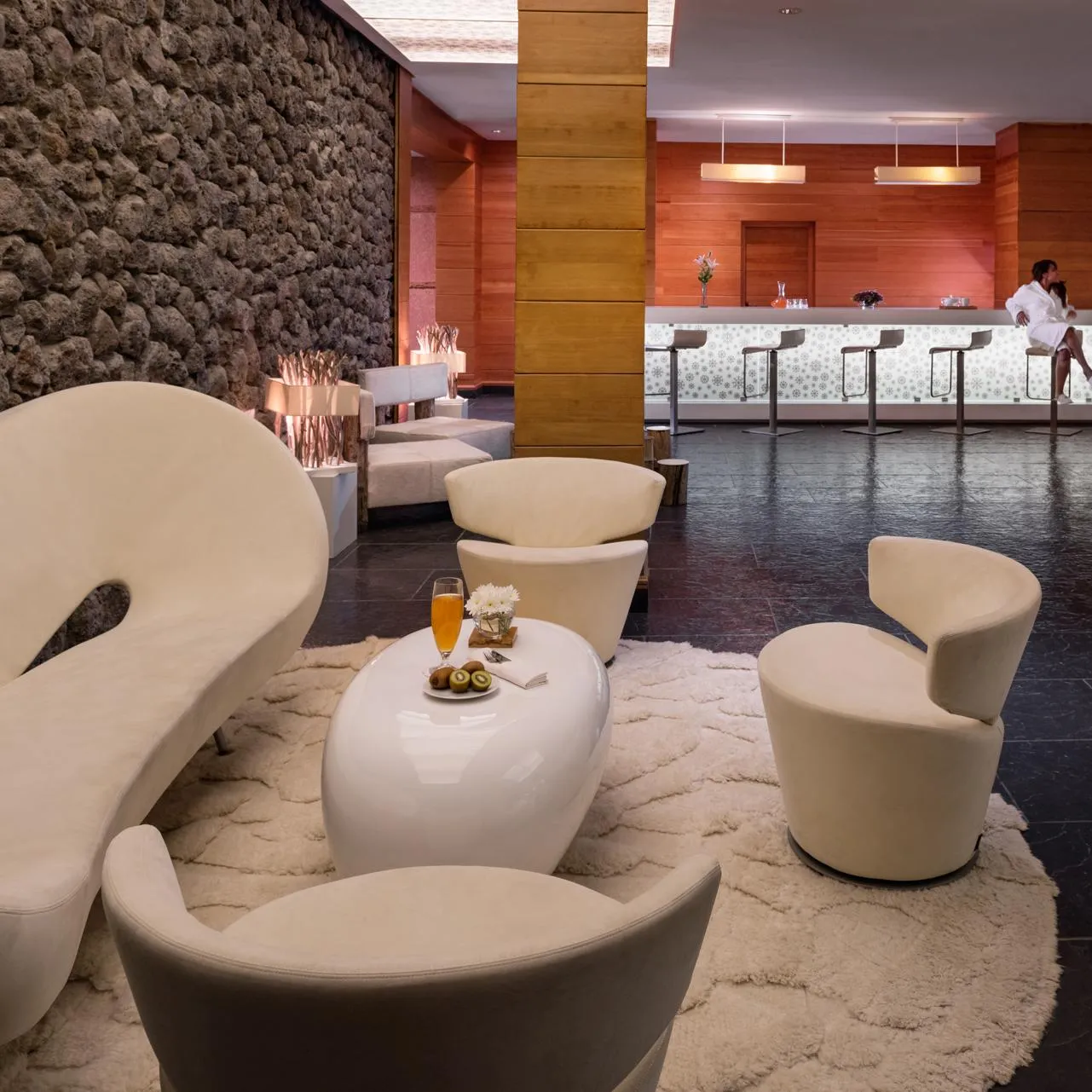 Un salon d'hôtel avec des sièges modernes, une table basse avec des boissons et des fruits, et un bar avec des tabourets en arrière-plan