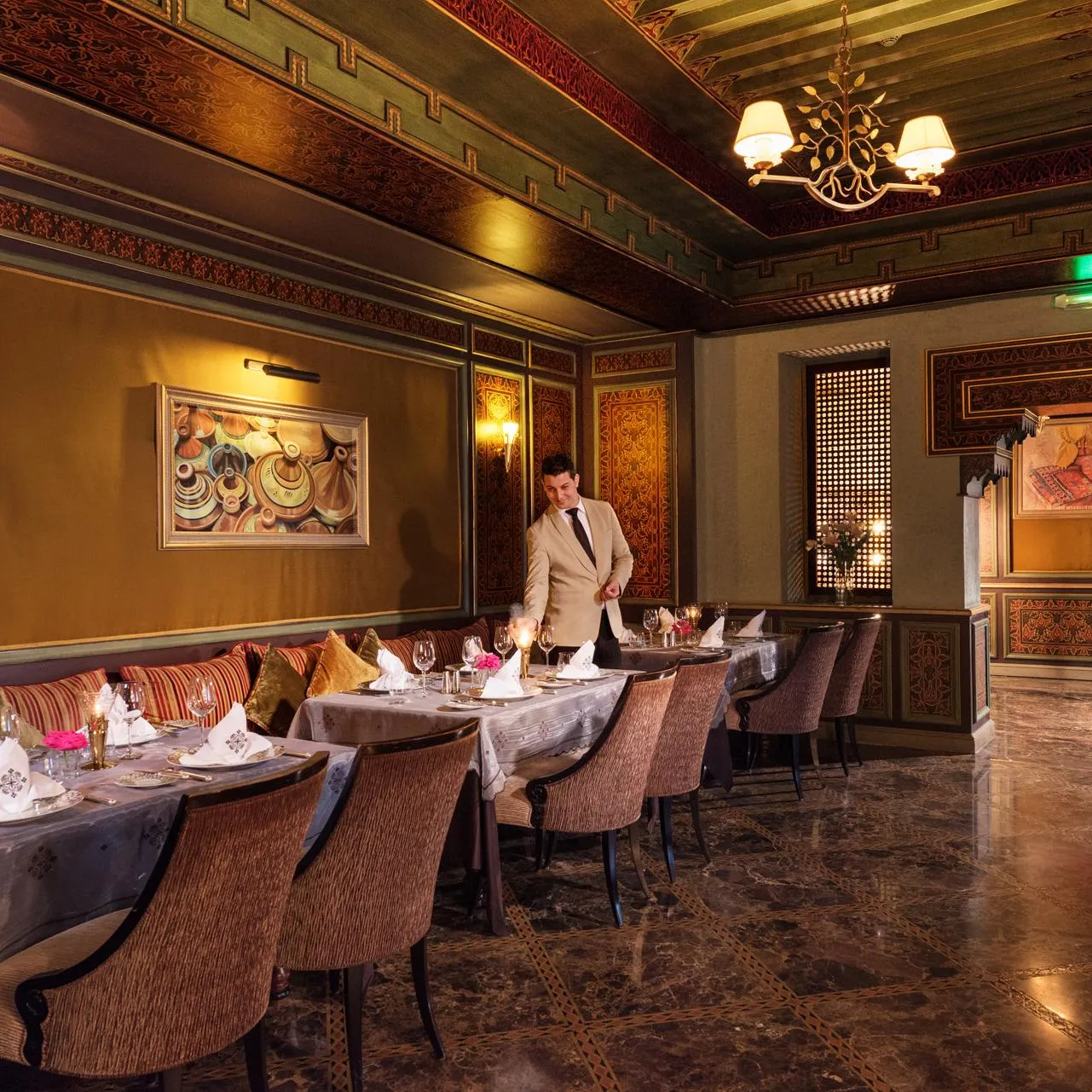 Un serveur en costume beige s'affaire dans un restaurant élégant aux murs ornés de motifs et de tableaux, avec des tables dressées pour le service