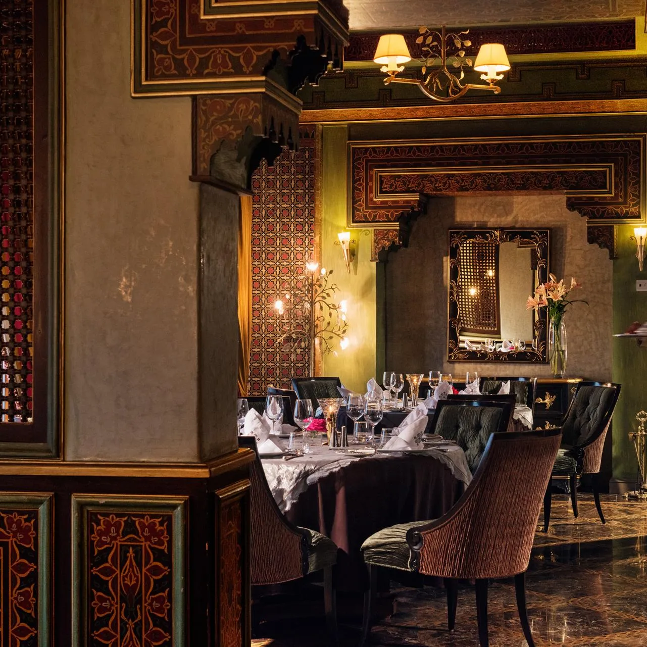 Un serveur en costume porte un plateau dans un restaurant élégant aux murs décorés de motifs et éclairé par des lampes murales