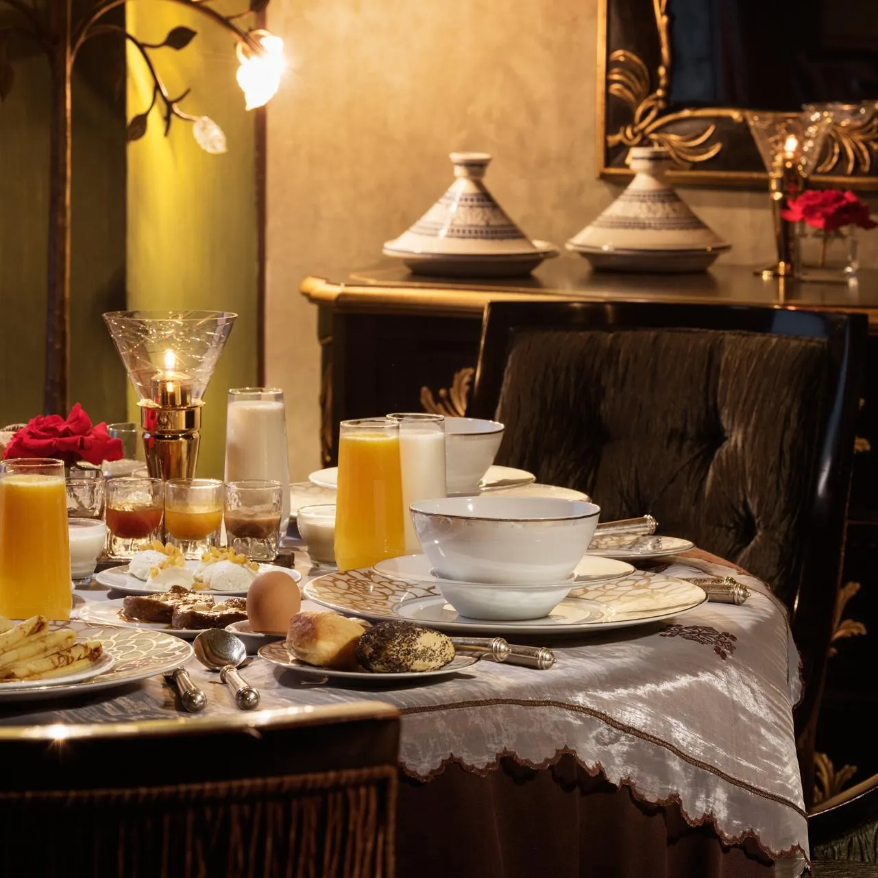 Table dressée pour le petit-déjeuner avec des boissons, des œufs, des viennoiseries et des fruits, dans un cadre élégant avec des chandeliers et des coussins