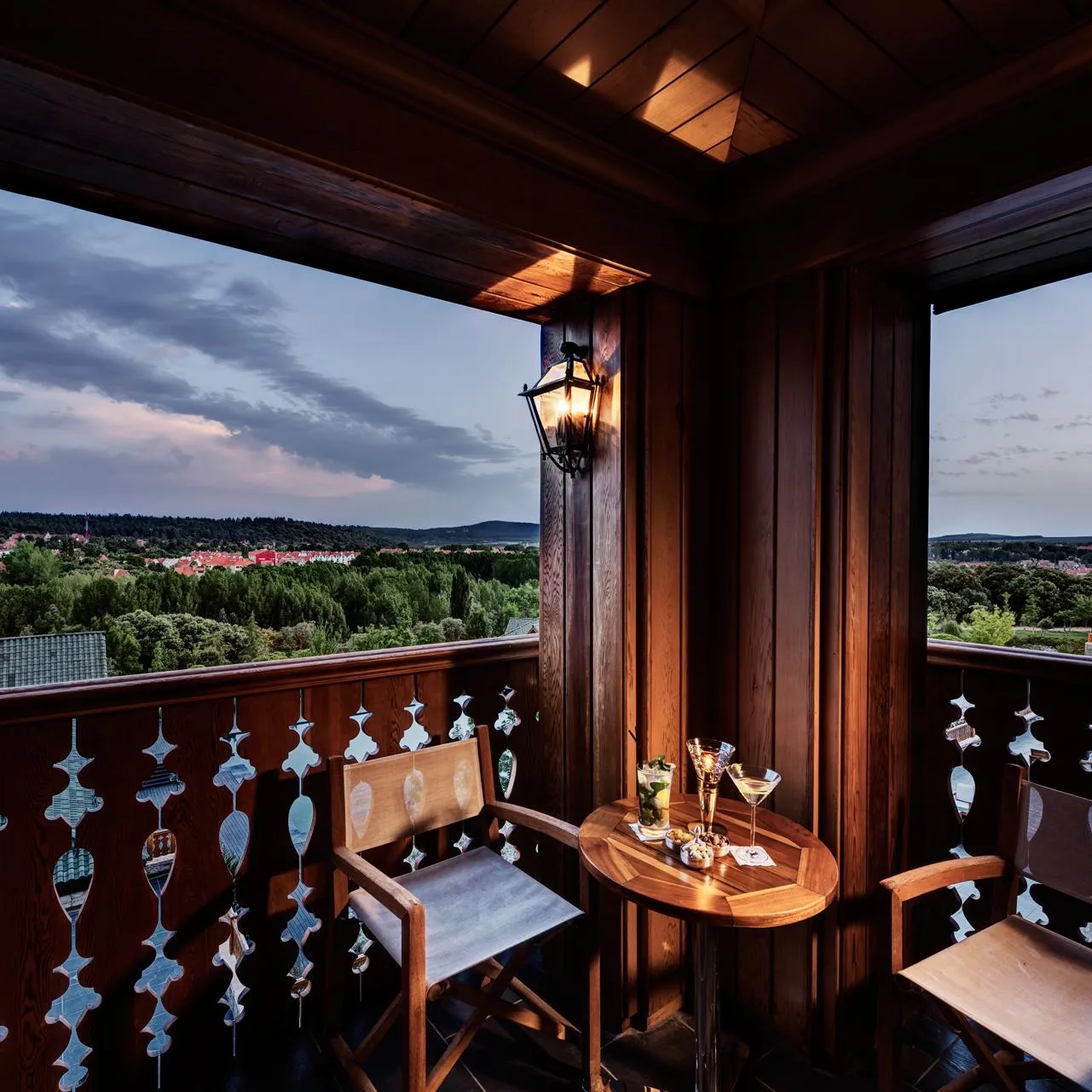 Balcon en bois avec table, chaises et verres, offrant une vue panoramique sur une ville et des collines sous un ciel nuageux