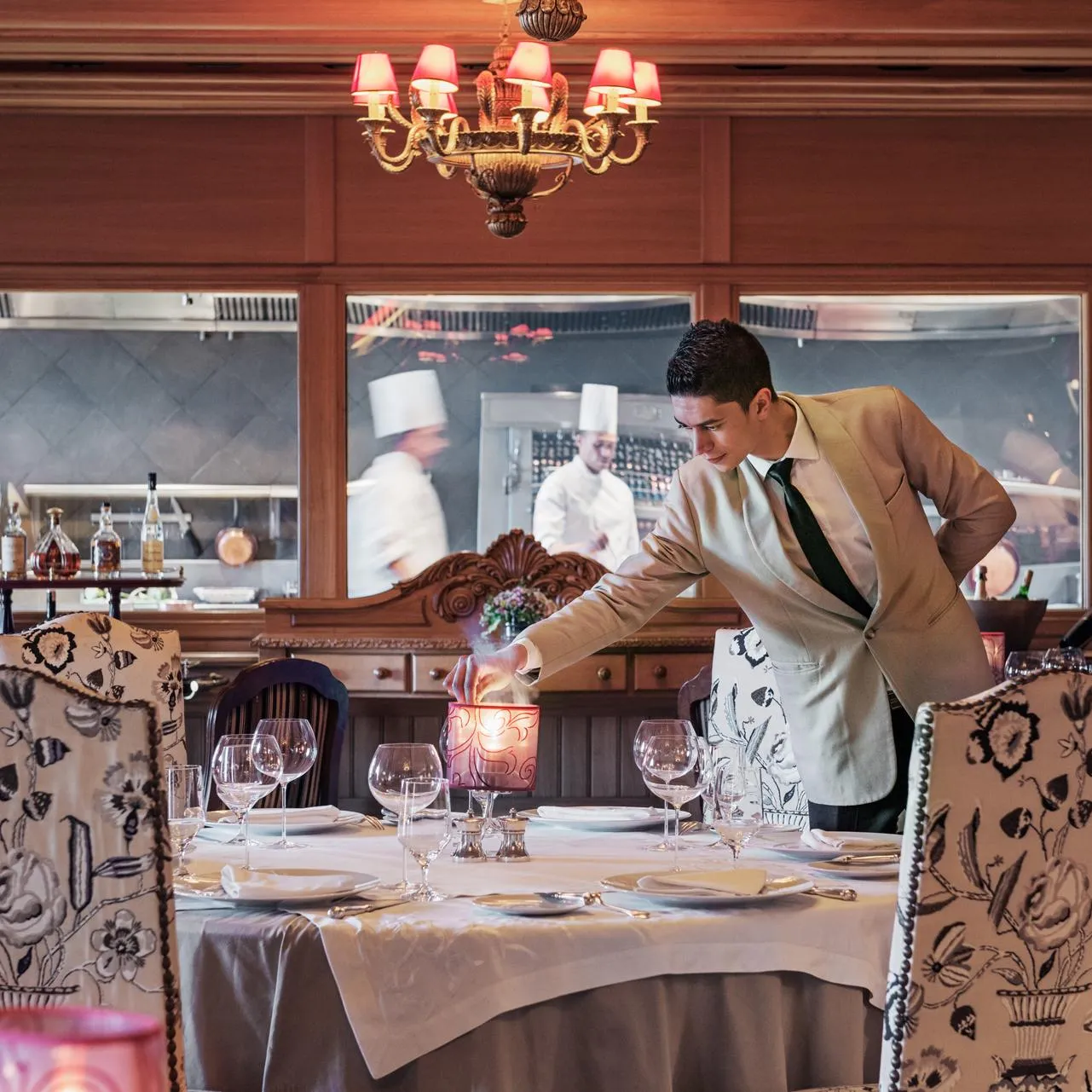 Un serveur en costume allume une bougie sur une table dressée dans un restaurant élégant avec une cuisine ouverte en arrière-plan