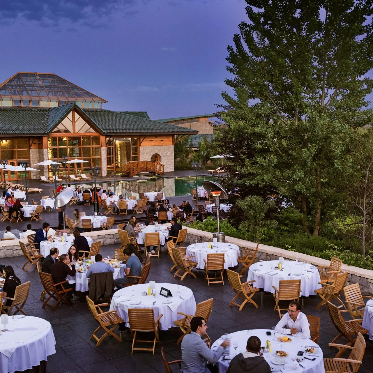 Une terrasse de restaurant en extérieur avec de nombreuses tables et chaises, des clients dînant et un bâtiment en bois et pierre en arrière-plan
