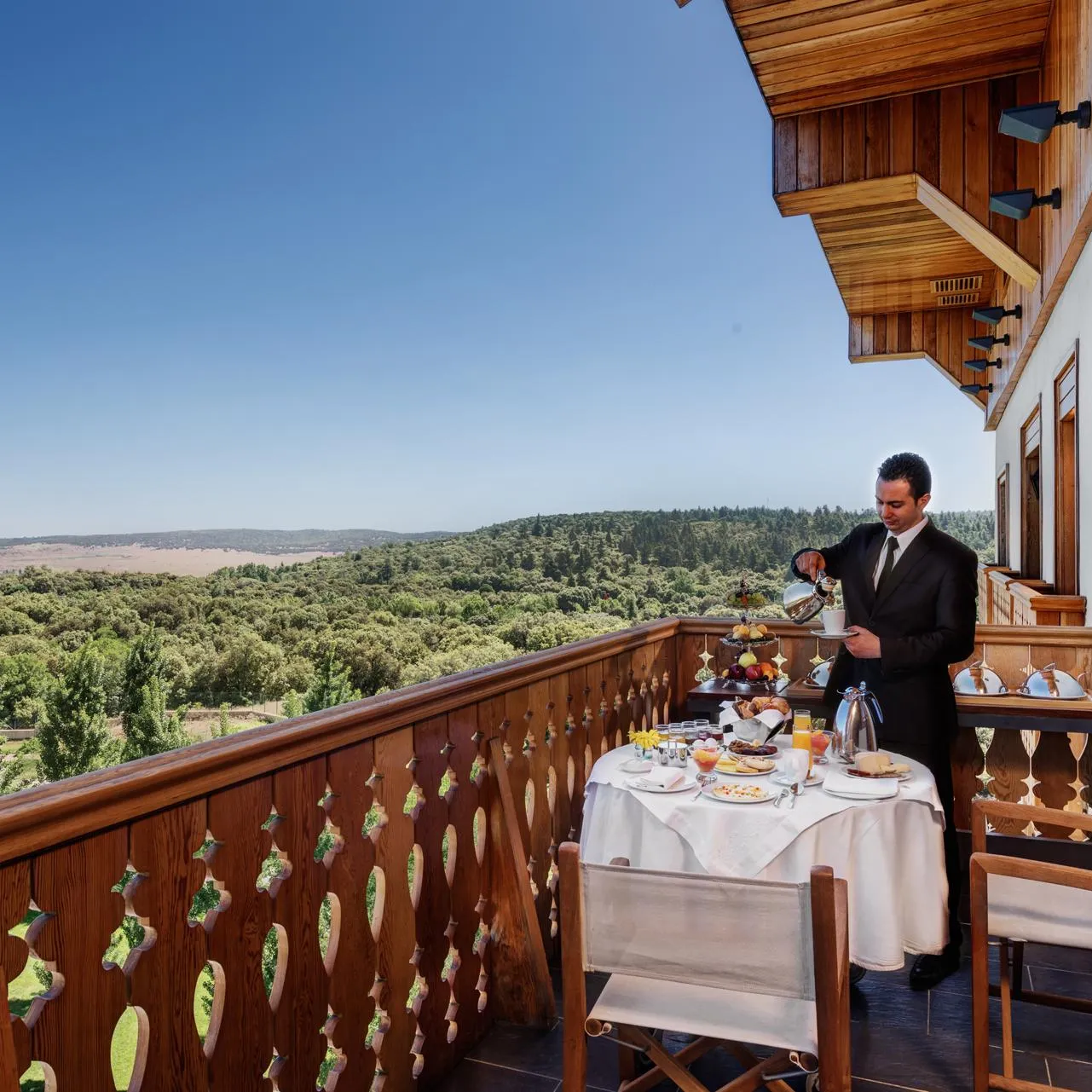 Un serveur en costume noir sert un repas sur une terrasse en bois avec vue sur une forêt et des collines