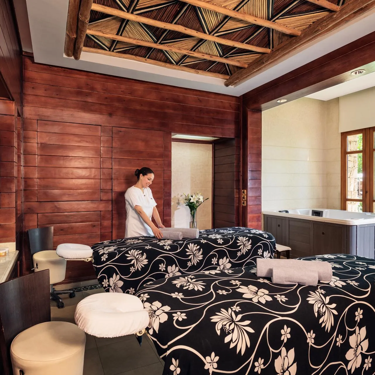 Une personne en tenue blanche prépare une table de massage dans une salle de spa en bois avec des fenêtres et des plantes