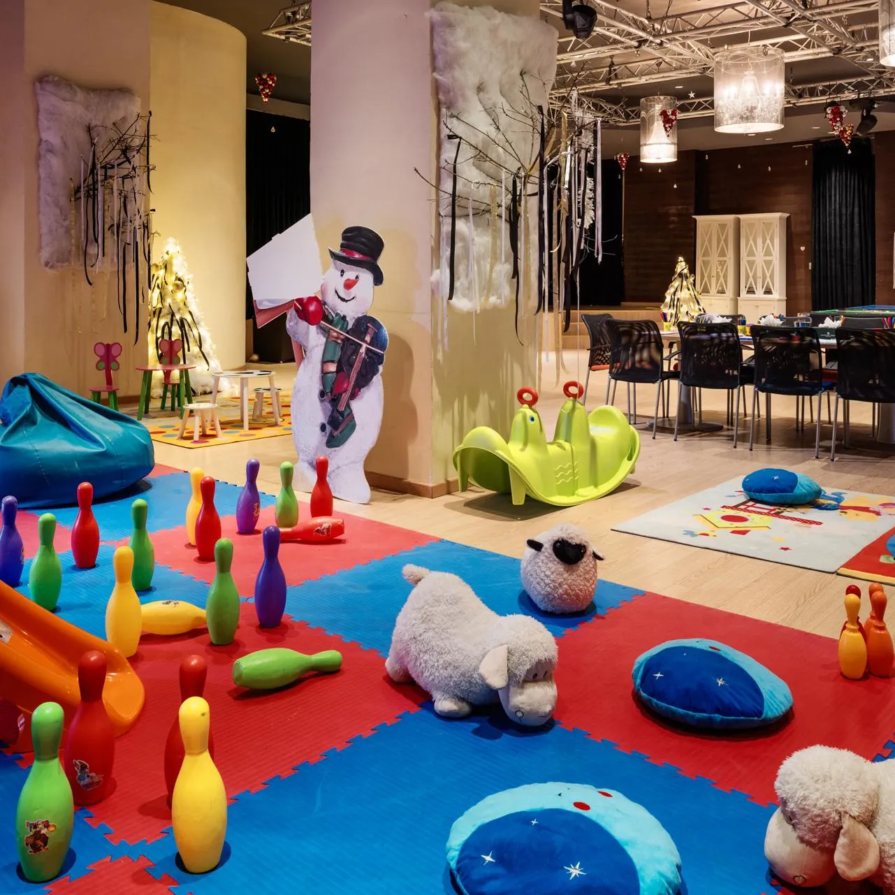 Salle de jeux pour enfants avec un snowman en carton, des quilles colorées, des coussins et des jouets sur des tapis rouges et bleus