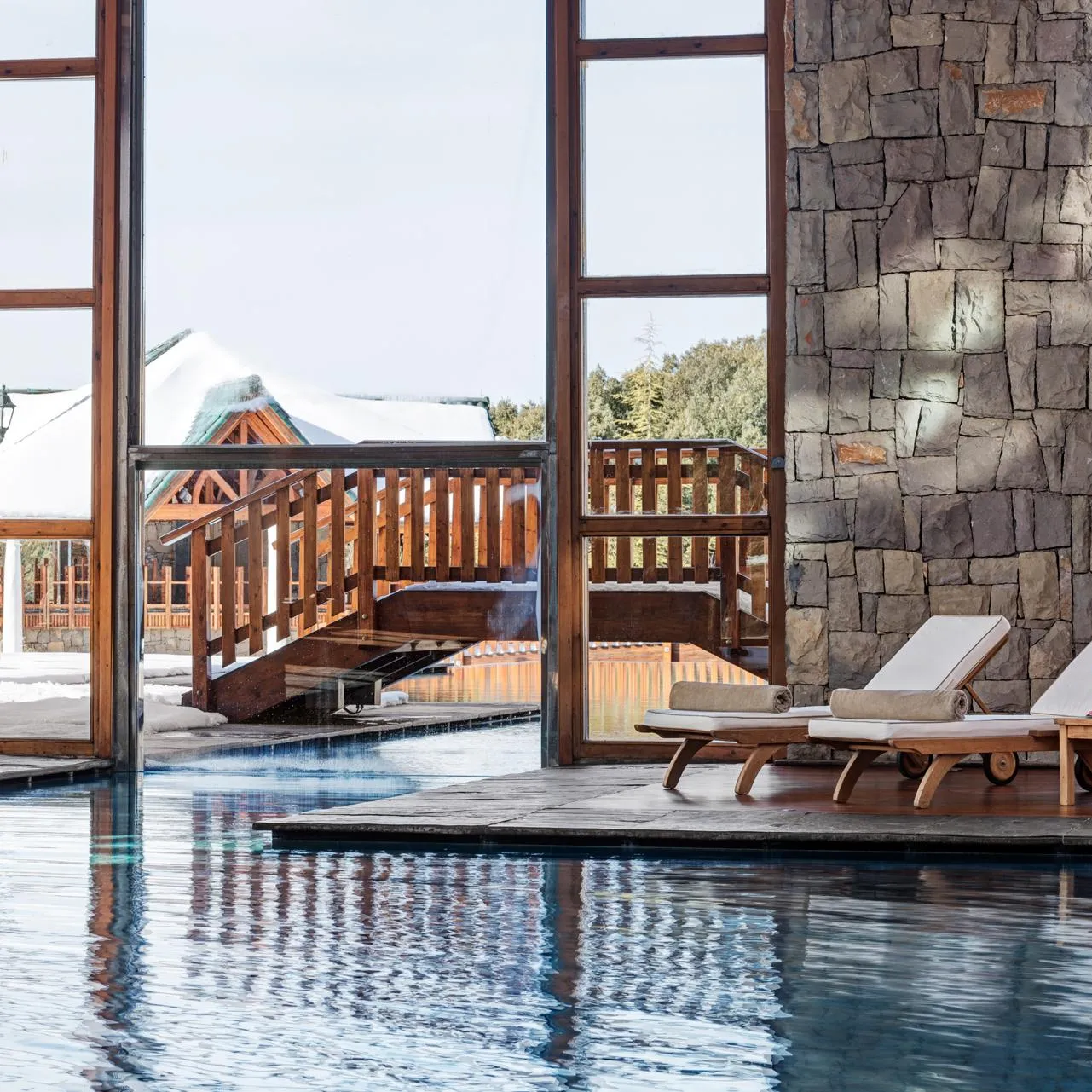 Deux transats avec serviettes et boissons sont placés près d'une piscine intérieure, avec une vue sur un extérieur enneigé à travers de grandes baies vitrées