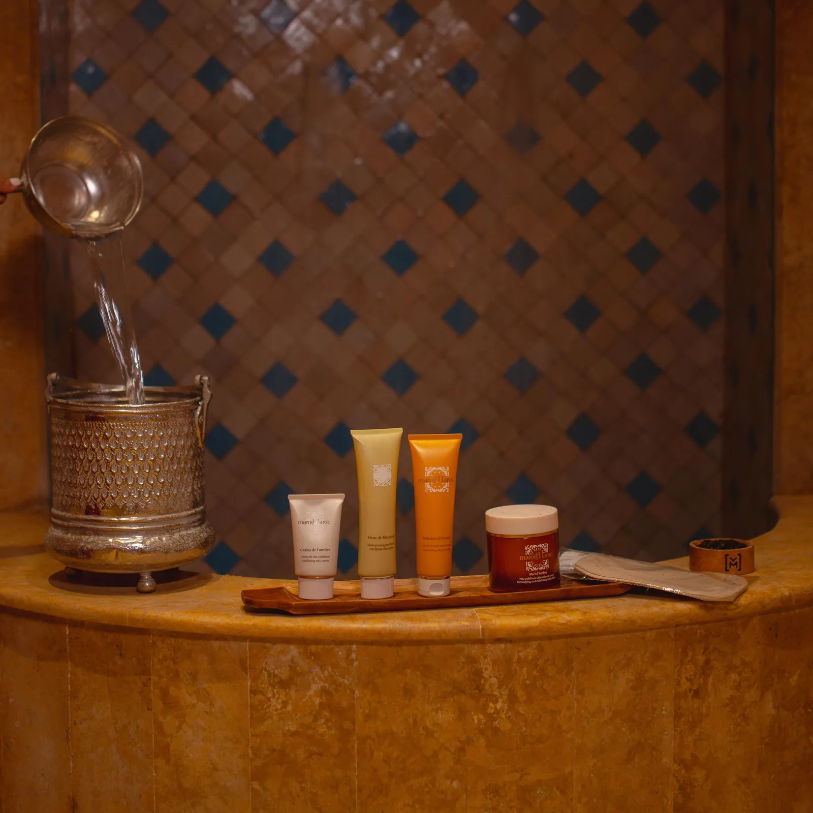 Un plateau en bois avec des tubes de produits cosmétiques, un pot et une spatule, à côté d'un récipient métallique avec un verseur, sur une étagère en pierre avec un mur carrelé en arrière-plan