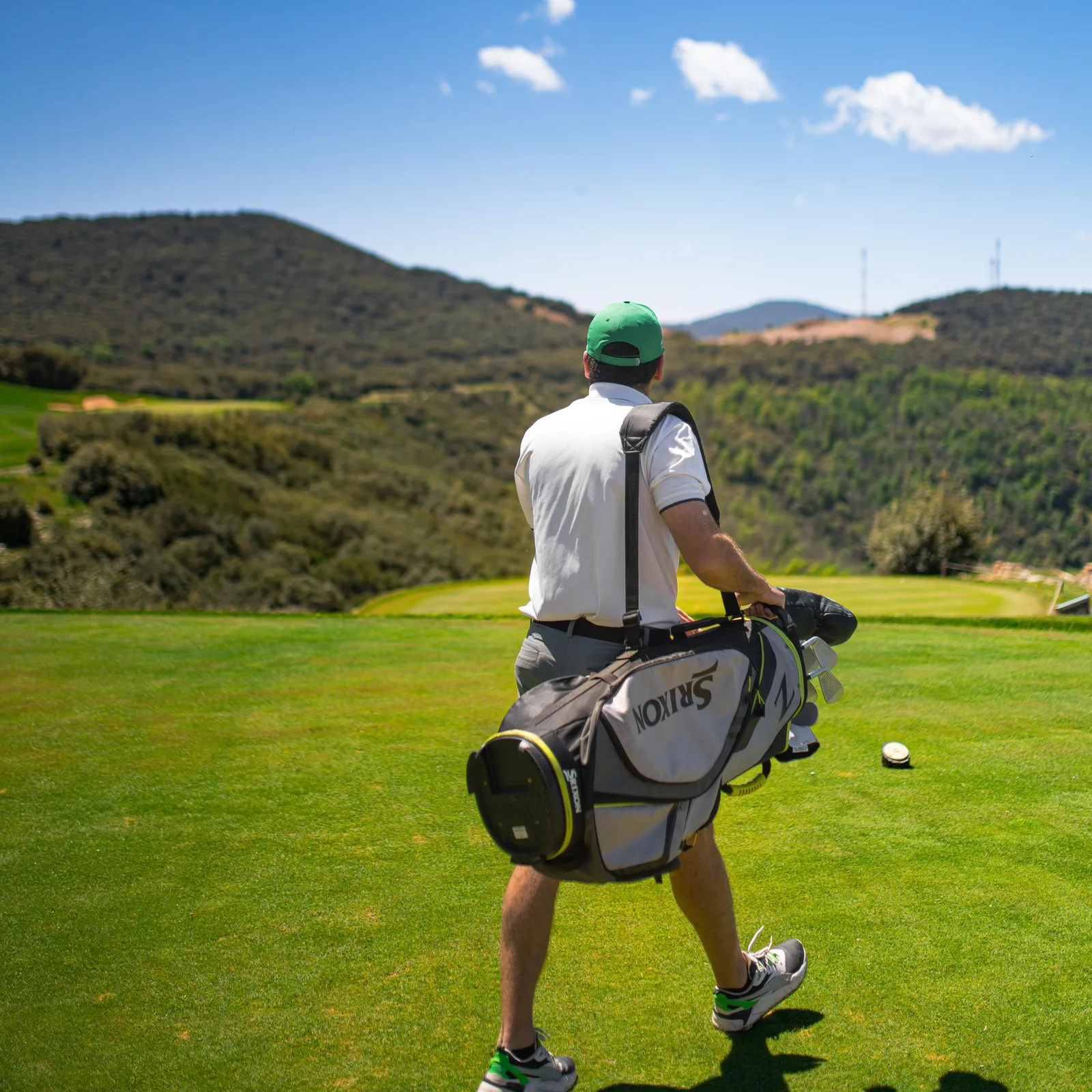 Un golfeur portant un sac de clubs marche sur un green avec des collines en arrière-plan