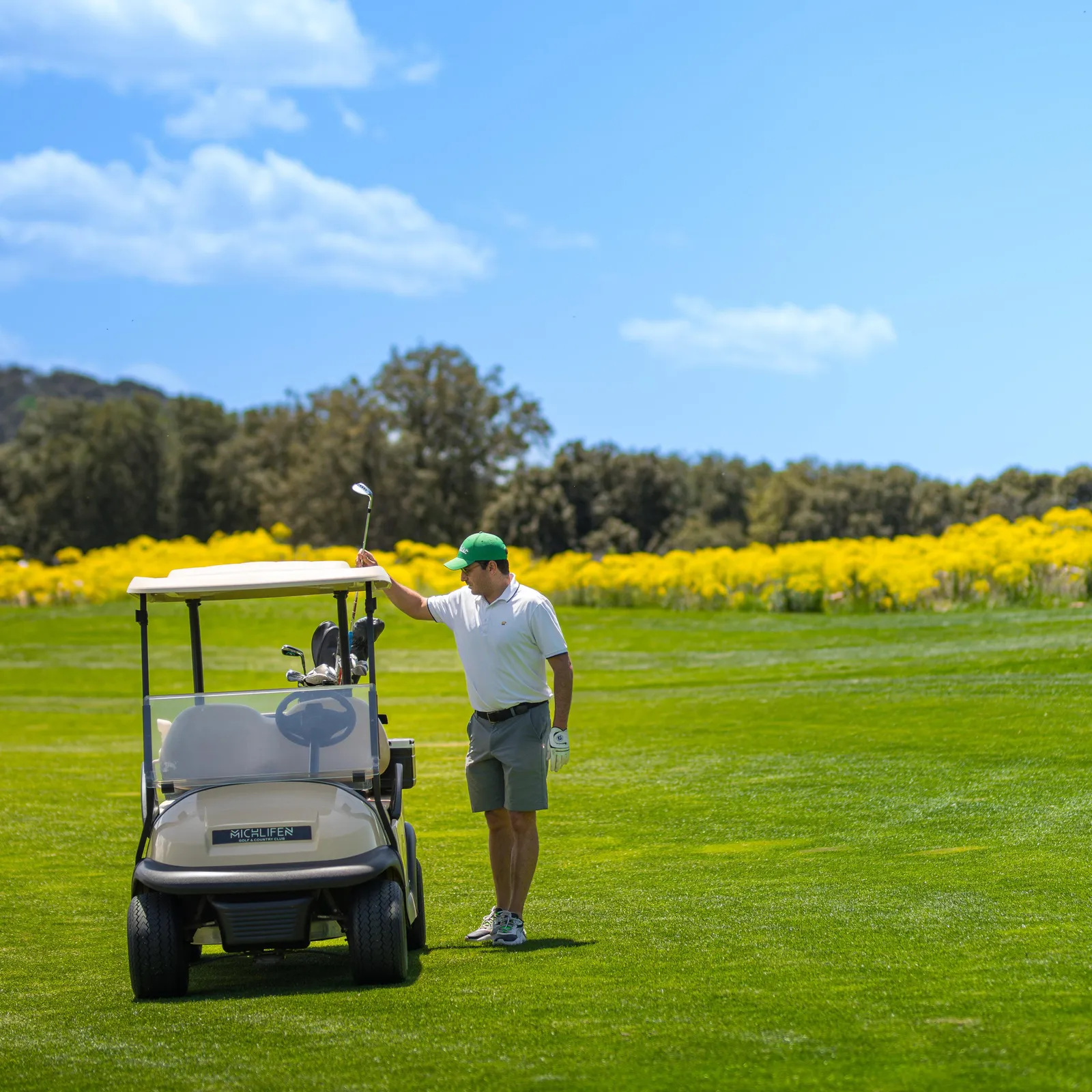 Un golfeur en tenue verte et blanche se tient près d'un chariot de golf sur un terrain de golf avec des fleurs jaunes en arrière-plan