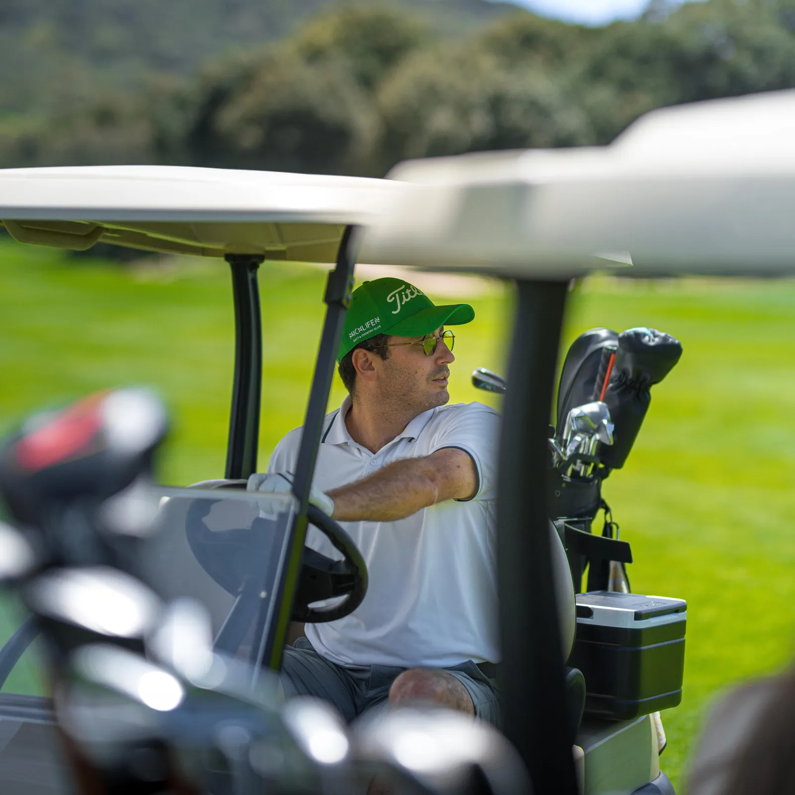 Un homme en chemise blanche et casquette verte conduit un véhicule de golf sur un parcours, avec des clubs de golf à l'arrière