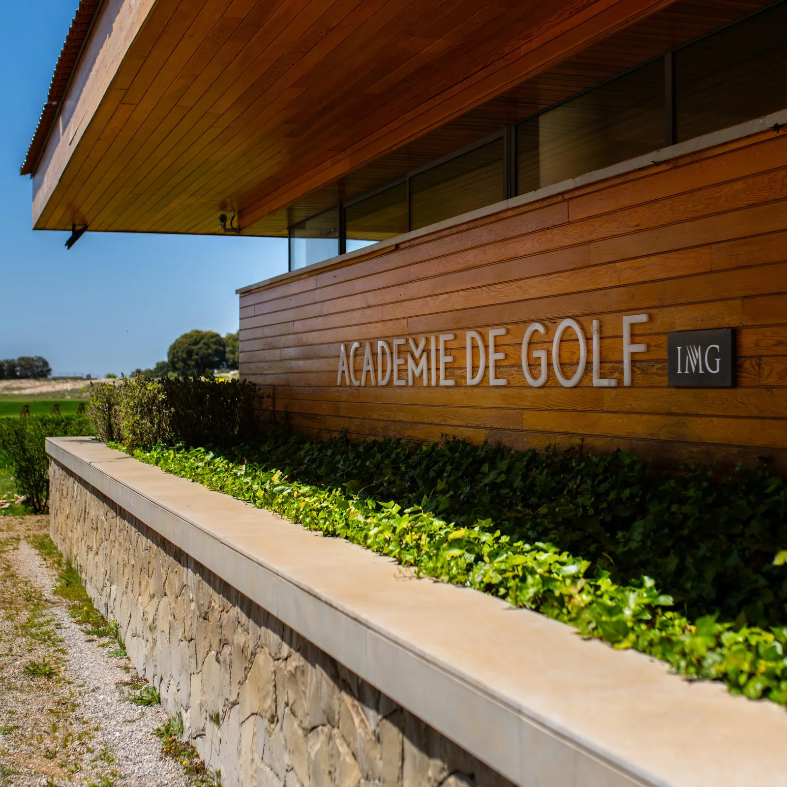 Bâtiment en bois avec l'inscription "Académie de Golf IMG" et un mur de pierre avec des plantes vertes
