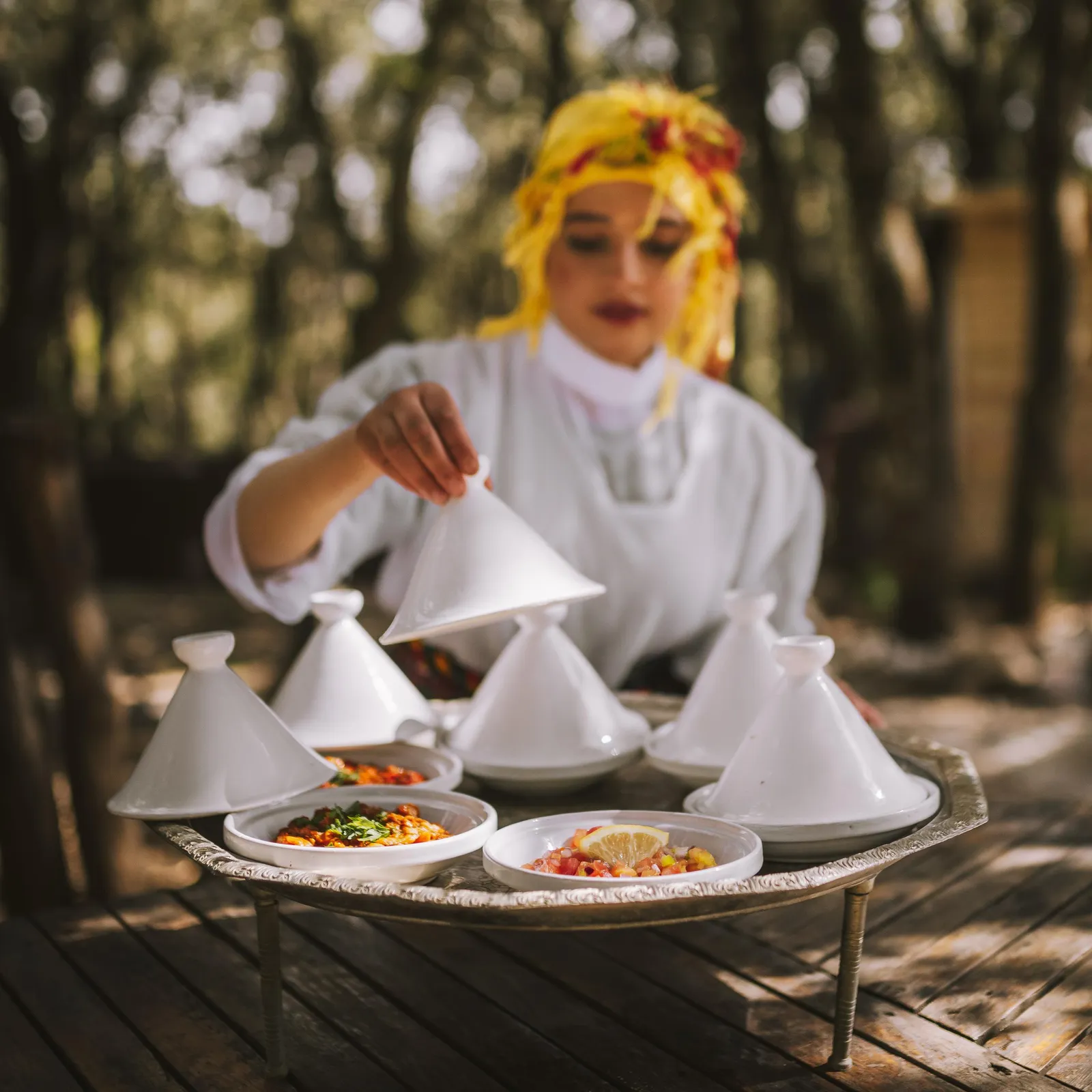 Une personne en tenue blanche et coiffe jaune prépare des plats dans des tajines posés sur un plateau métallique sur une table en bois en extérieur