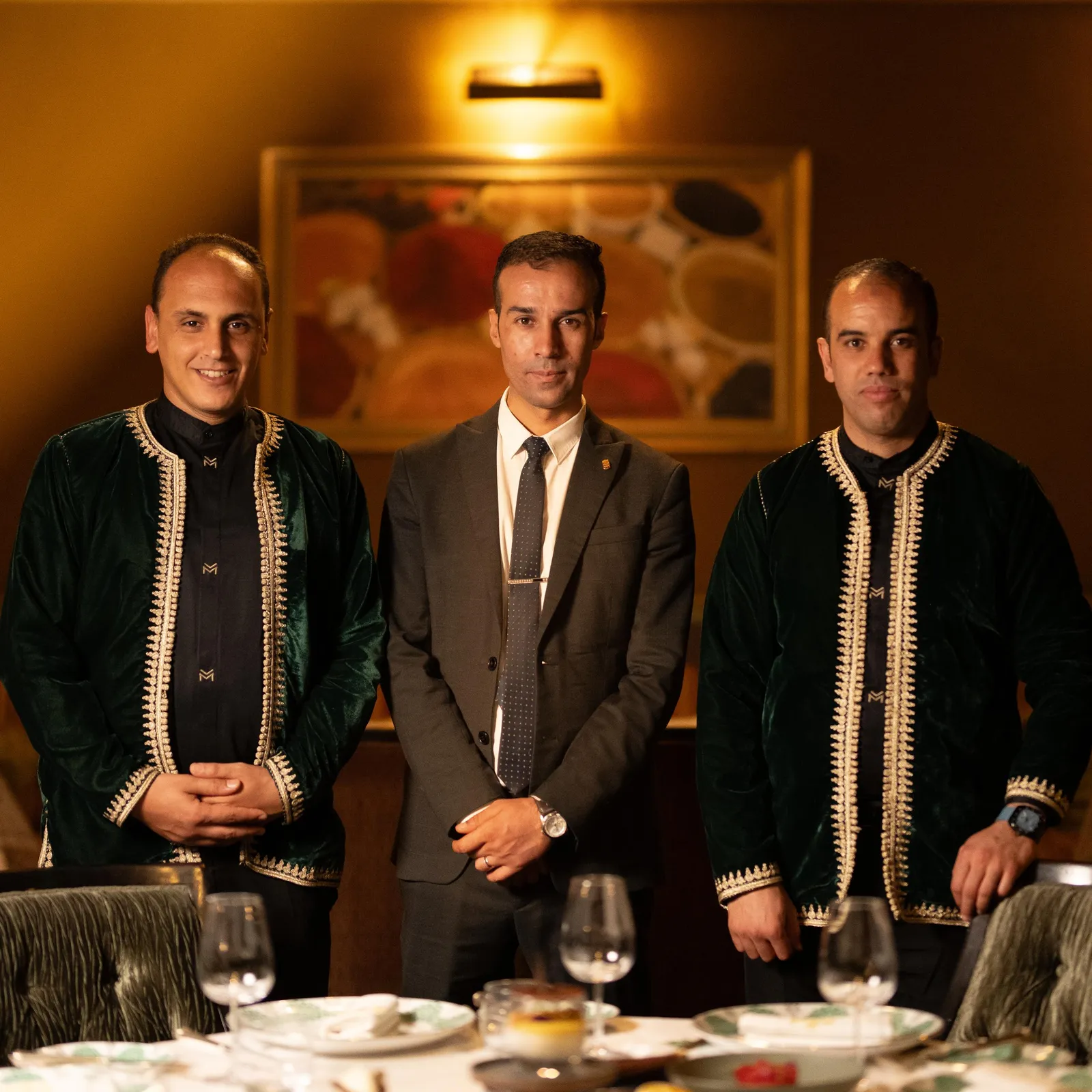 Trois hommes assis à une table avec des verres de vin, deux portant des vestes ornées et un en costume