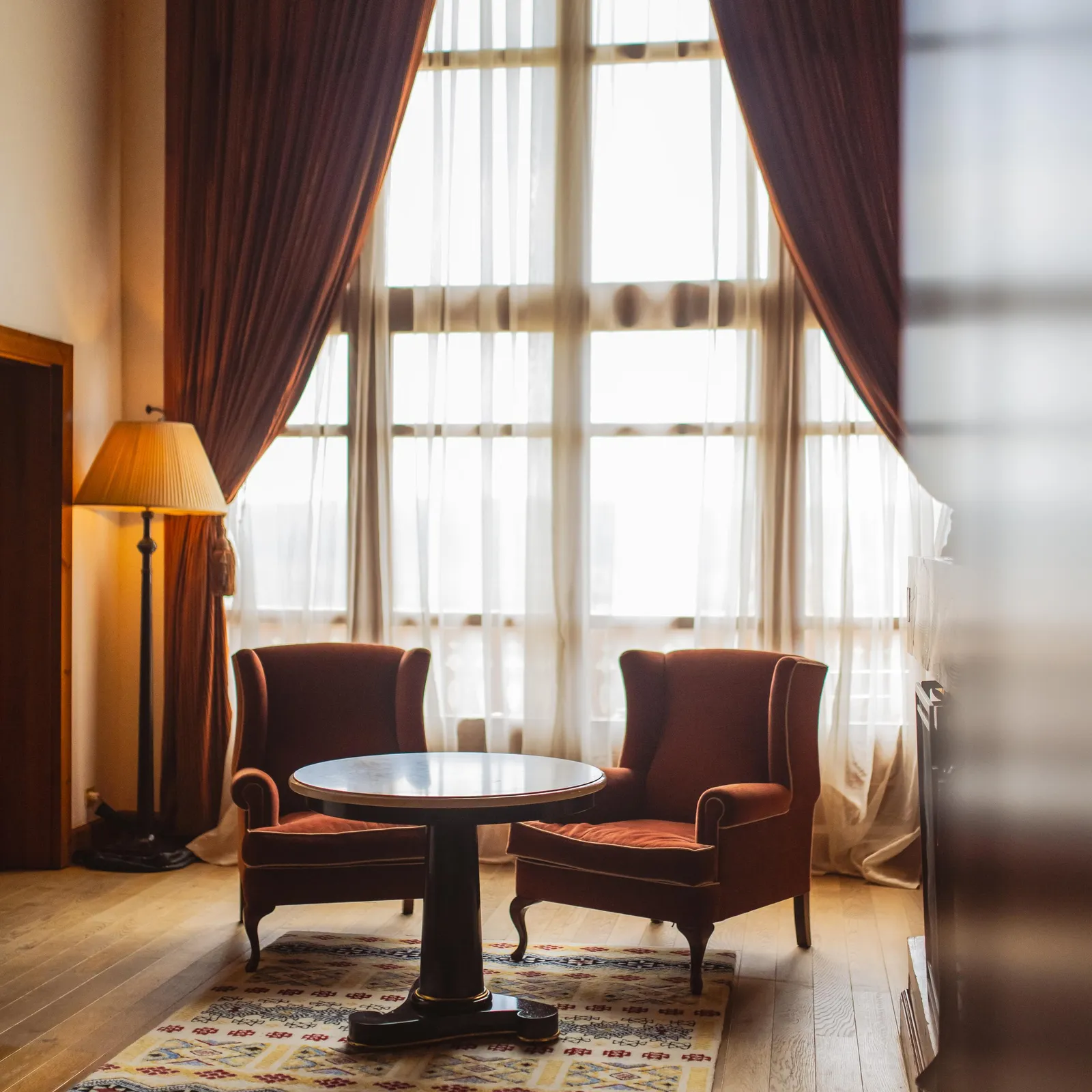 Deux fauteuils en velours rouge entourent une table ronde dans un salon éclairé par de grandes fenêtres aux rideaux marron